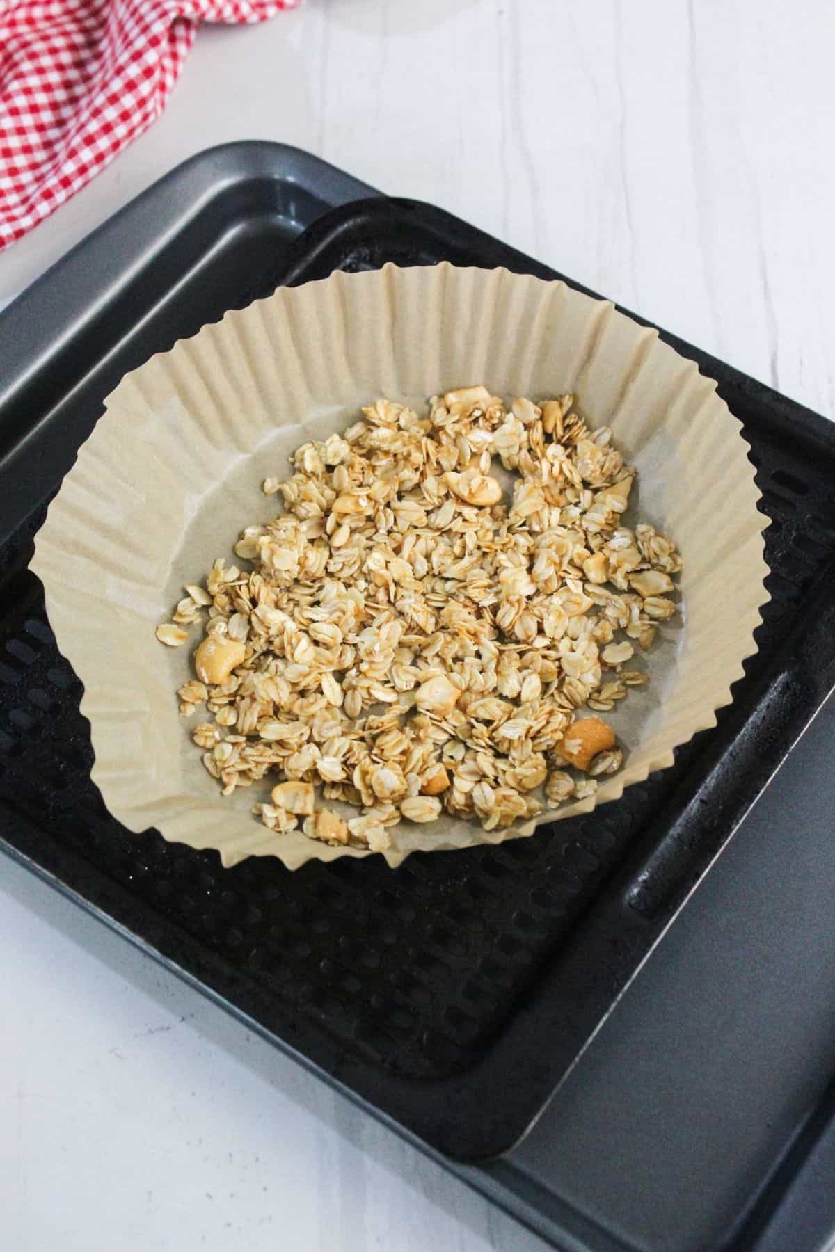 A baking tray with a large parchment paper liner holds a mixture of oats and nuts, ready to be baked. A red and white checkered cloth is in the background.