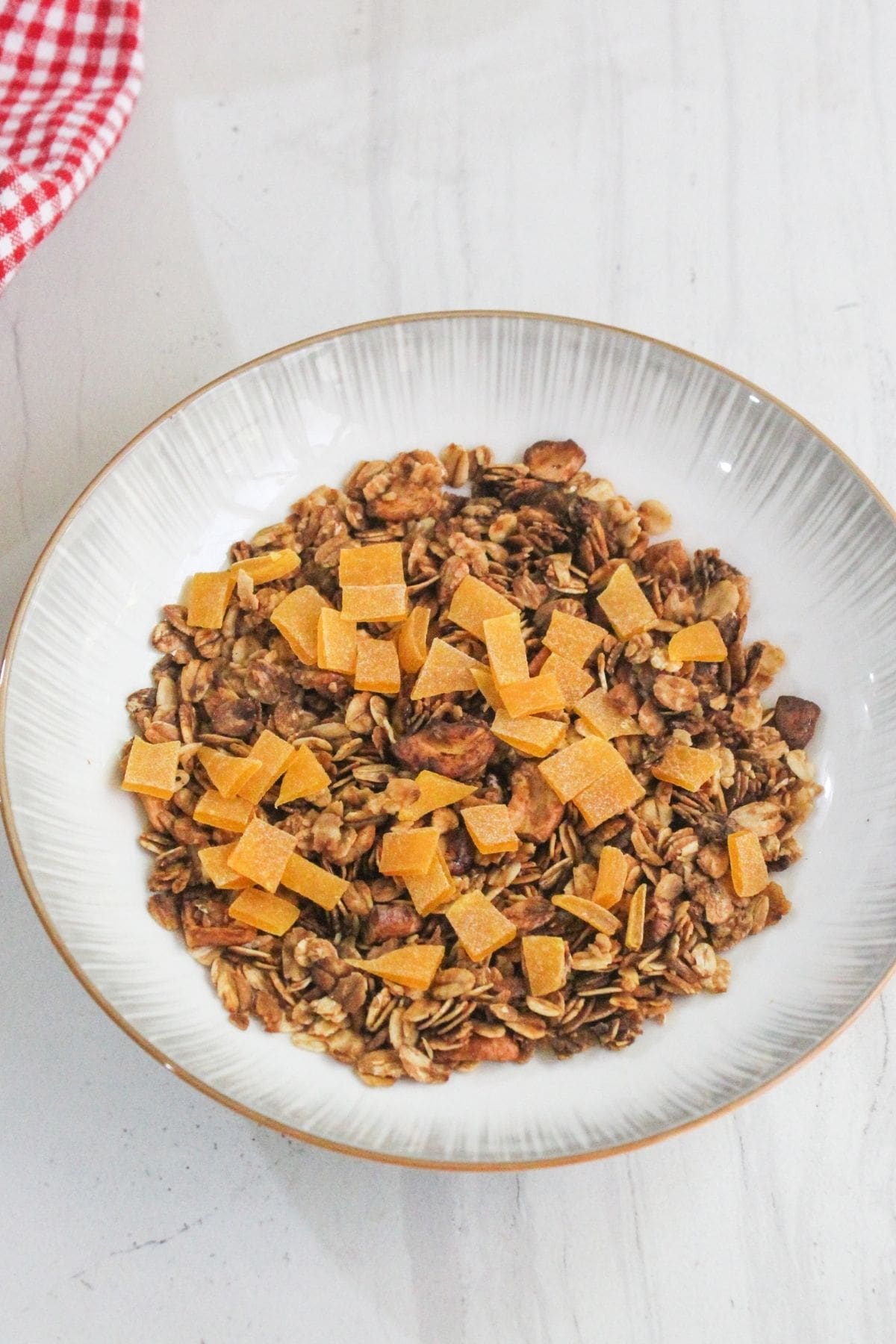 A bowl of granola topped with small square pieces of dried fruit, placed on a light-colored surface.