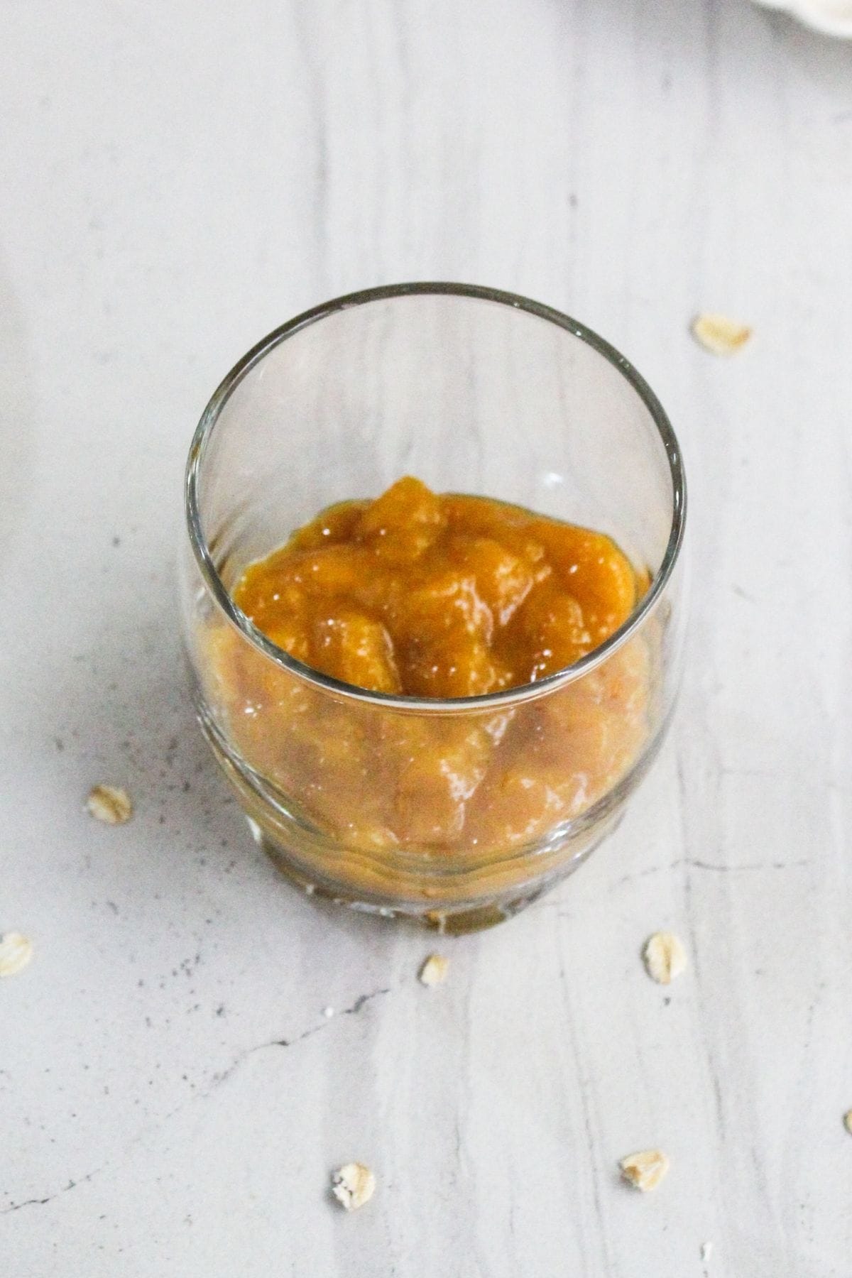 A clear glass filled with a thick, orange-brown sauce sits on a light surface with a few scattered oats nearby.