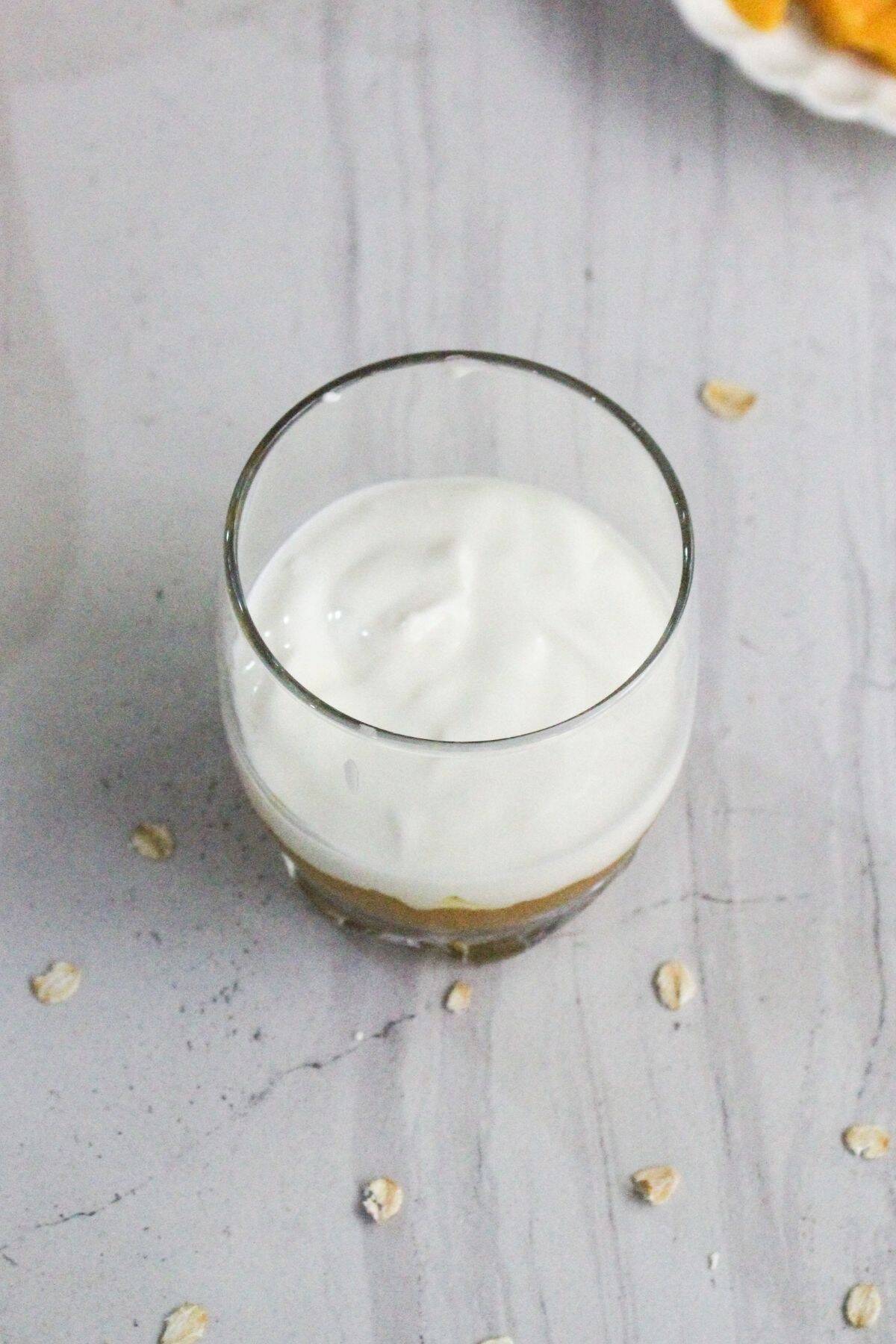 A glass filled with a layer of yogurt on top of what appears to be a fruit or oat mixture, placed on a light gray surface with scattered oat flakes around it.