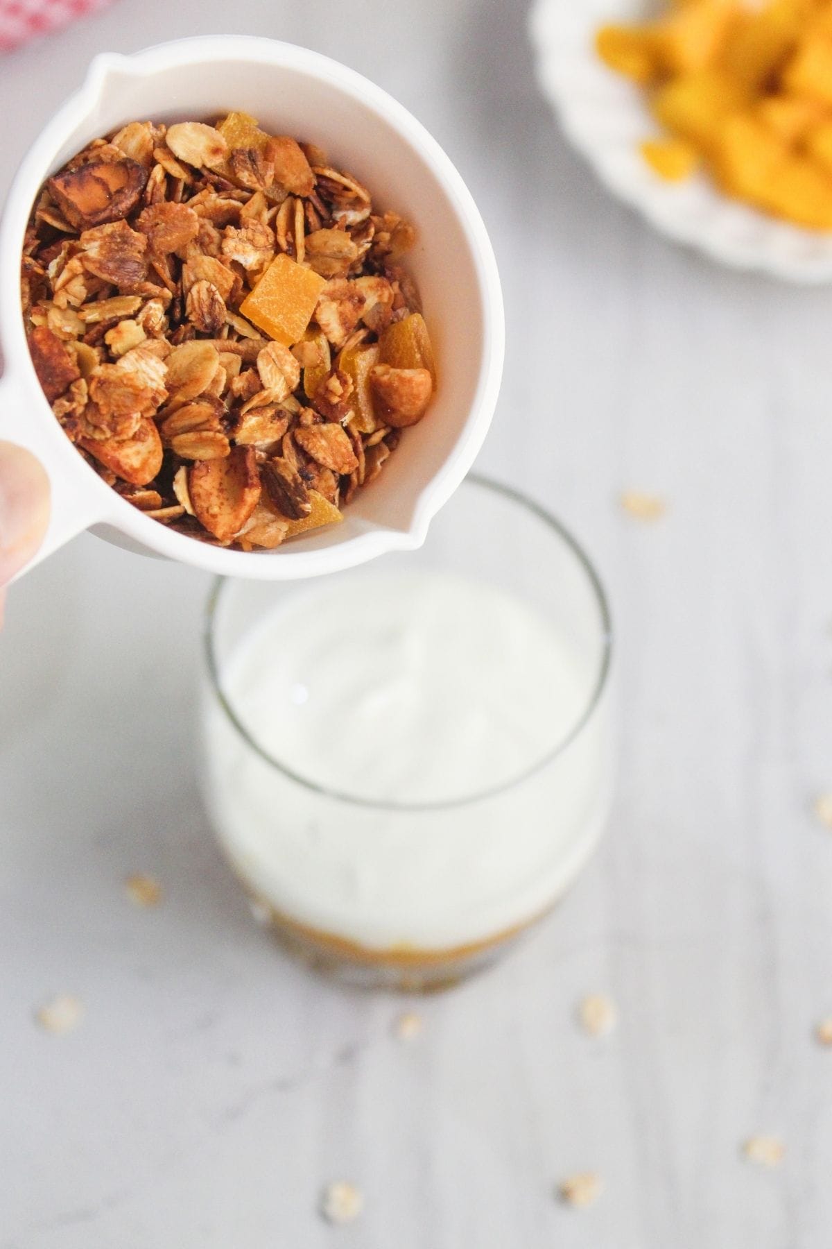 A hand pours granola from a white measuring cup into a glass containing yogurt, with a plate of chopped fruit in the background.