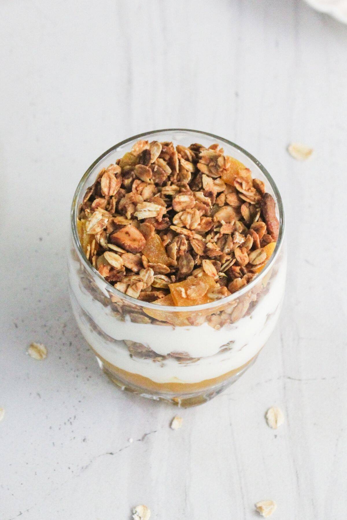 A glass cup filled with layered granola, yogurt, and fruit, placed on a light-colored surface with a few oats scattered around.