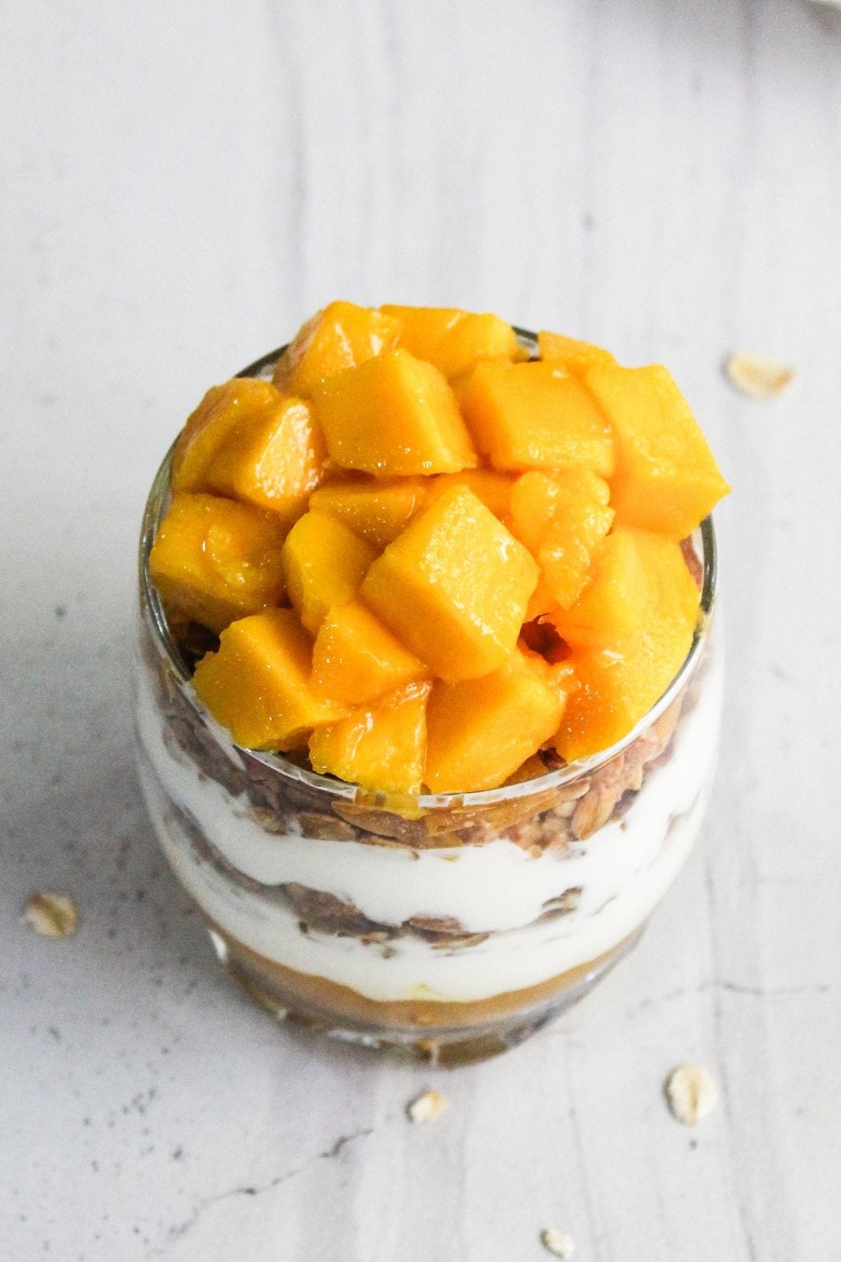 A glass cup filled with layers of yogurt, granola, and mango chunks, placed on a light-colored surface.