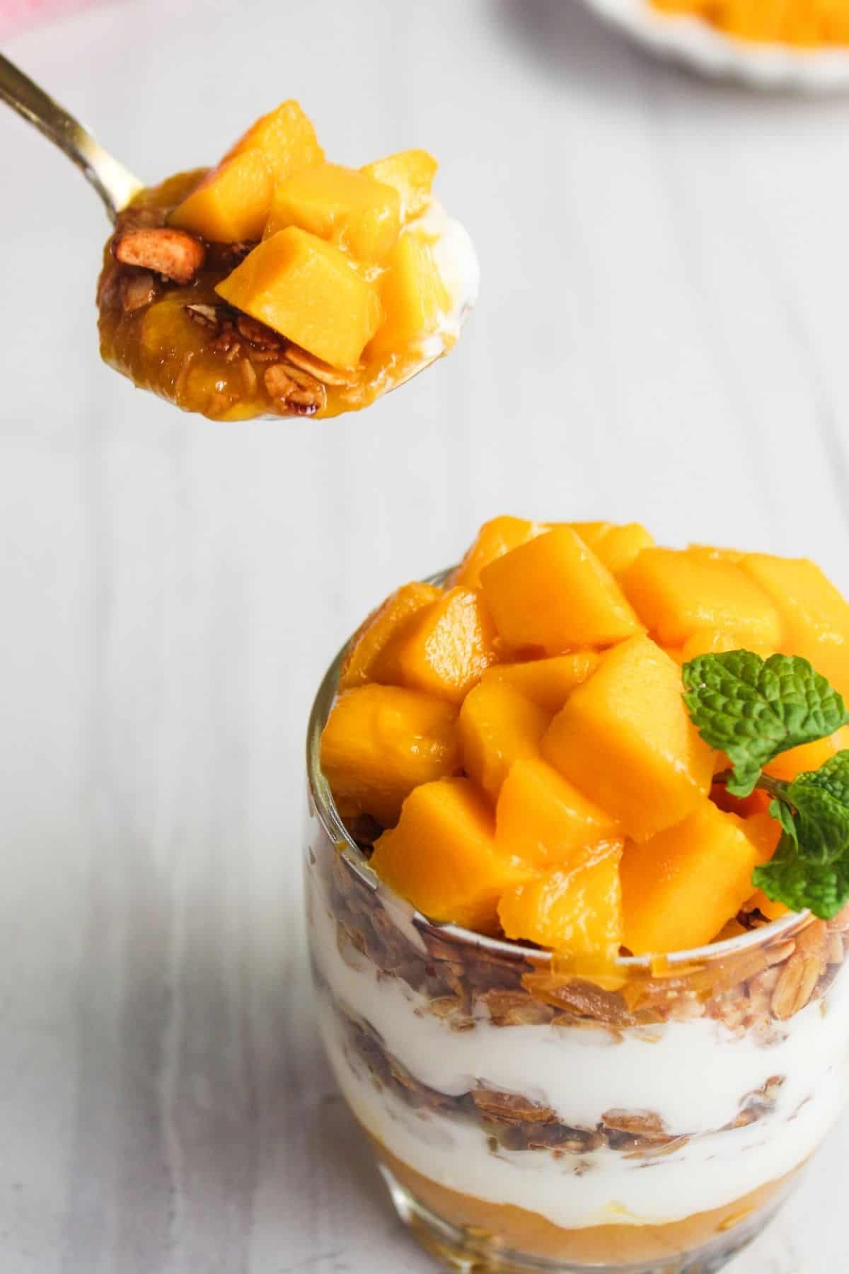 A glass of parfait layered with granola, yogurt, and diced mango, topped with mint, with a spoonful of parfait being held above.