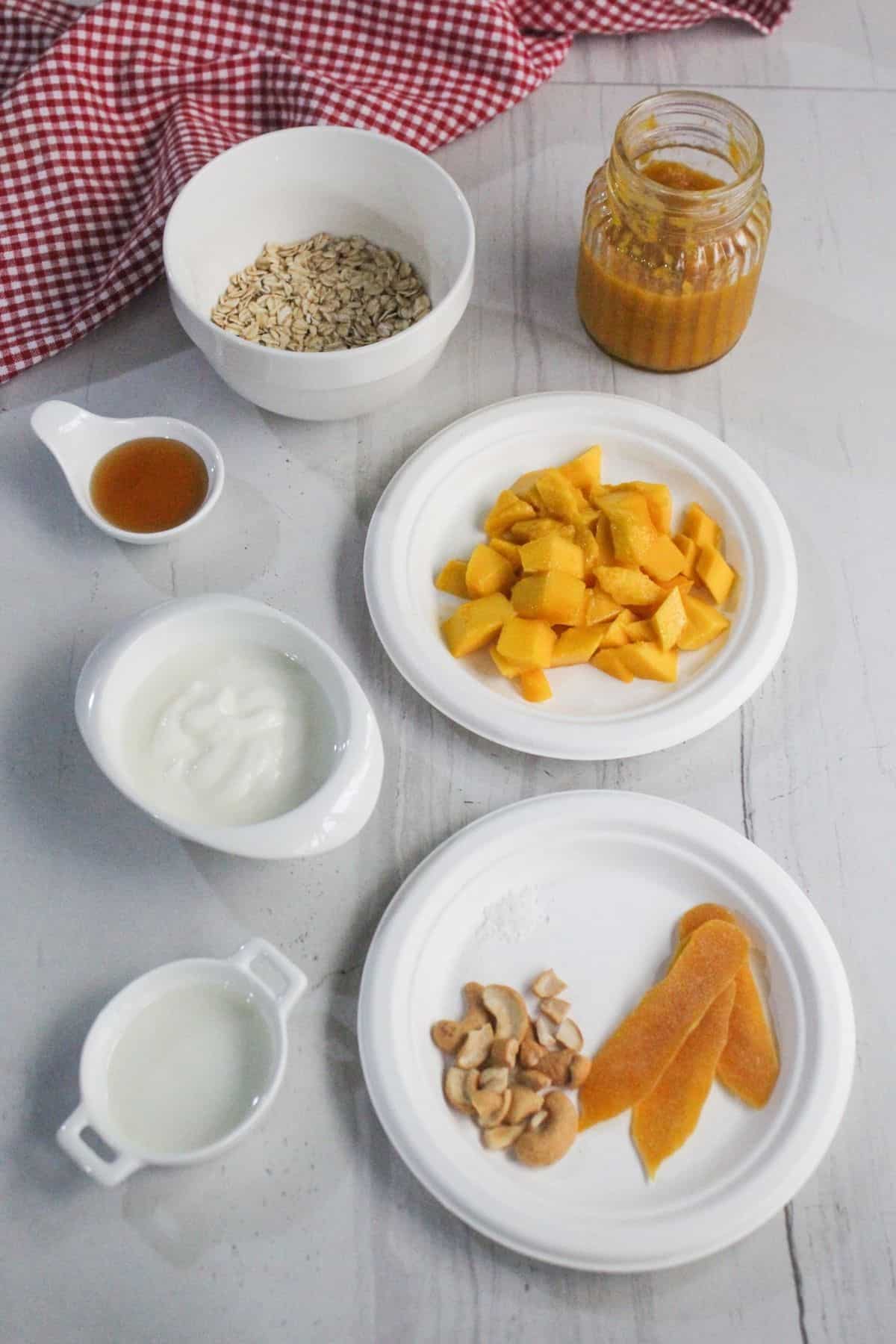 A flat lay of ingredients for a recipe, including oats, honey, yogurt, milk, chopped mango, dried mango slices, cashews, and a jar of orange sauce on a white surface.