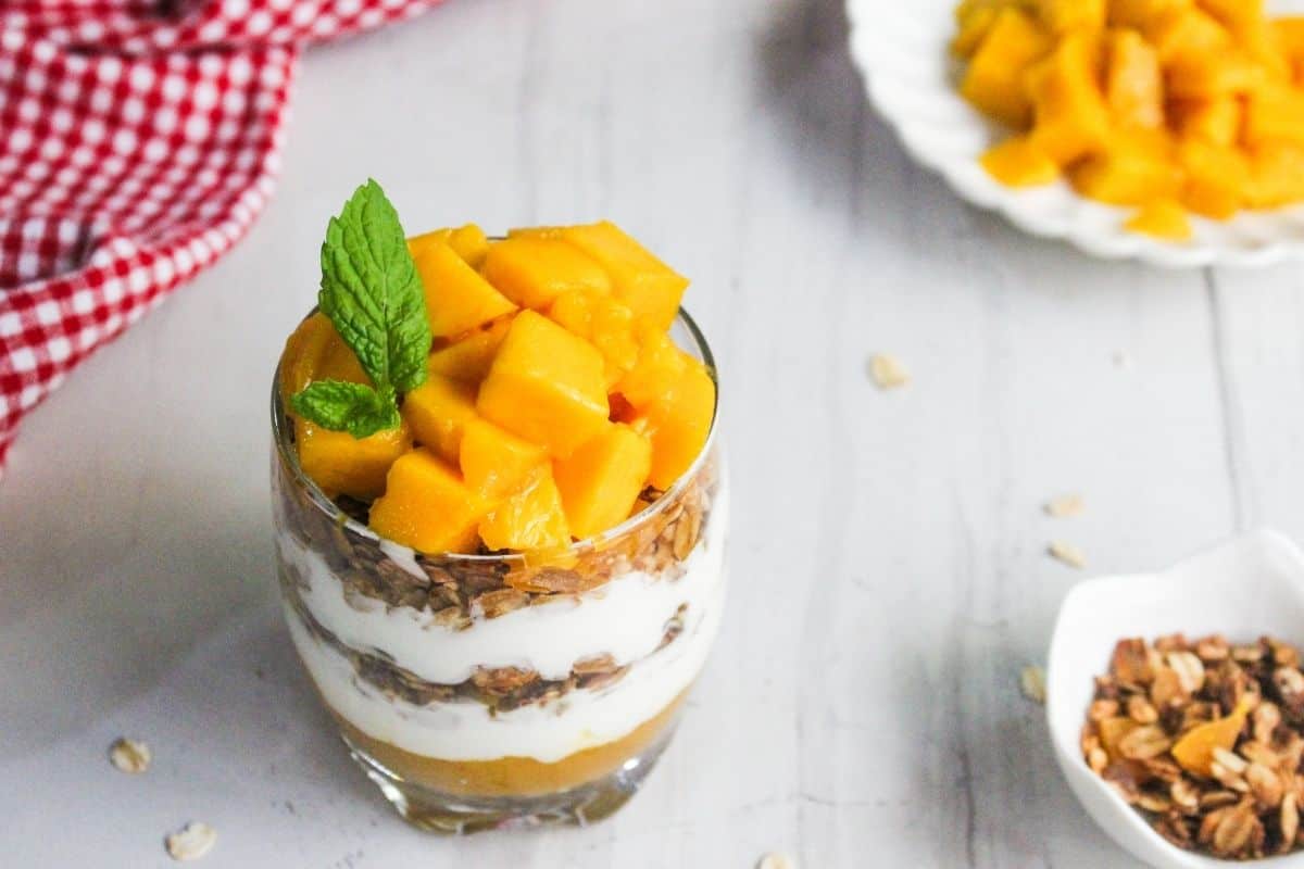 A glass filled with layers of granola, yogurt, and mango chunks, garnished with a mint leaf. Granola and diced mango are also placed in bowls nearby.