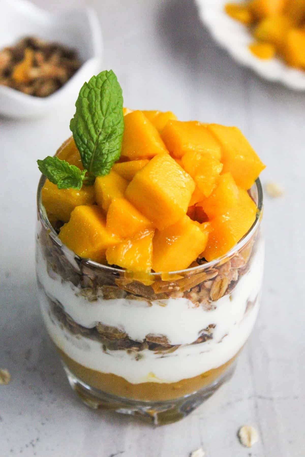 A glass parfait with layers of granola, yogurt, and mango chunks, topped with fresh mint.
