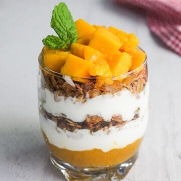 A glass filled with layers of mango puree, granola, and yogurt, topped with diced mango and a mint sprig.
