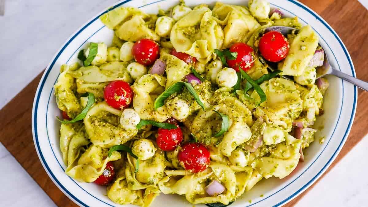 A bowl of tortellini pasta salad with pesto, cherry tomatoes, mozzarella balls, red onion, and fresh basil, served with a spoon.