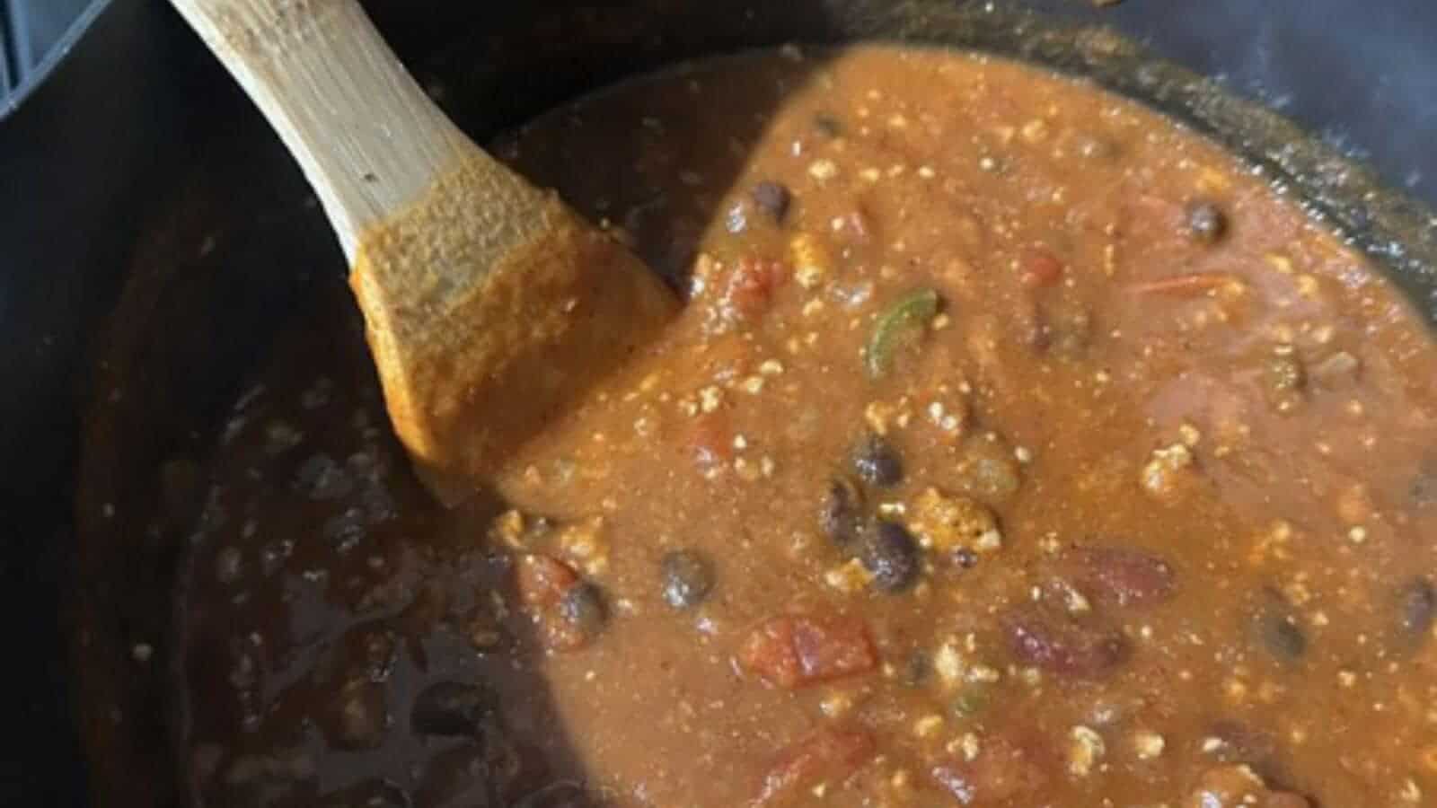 A pot of chili with beans, ground meat, and tomatoes being stirred with a wooden spoon—a cozy dish that’s perfect for autumn and pairs perfectly with your favorite pumpkin recipes.