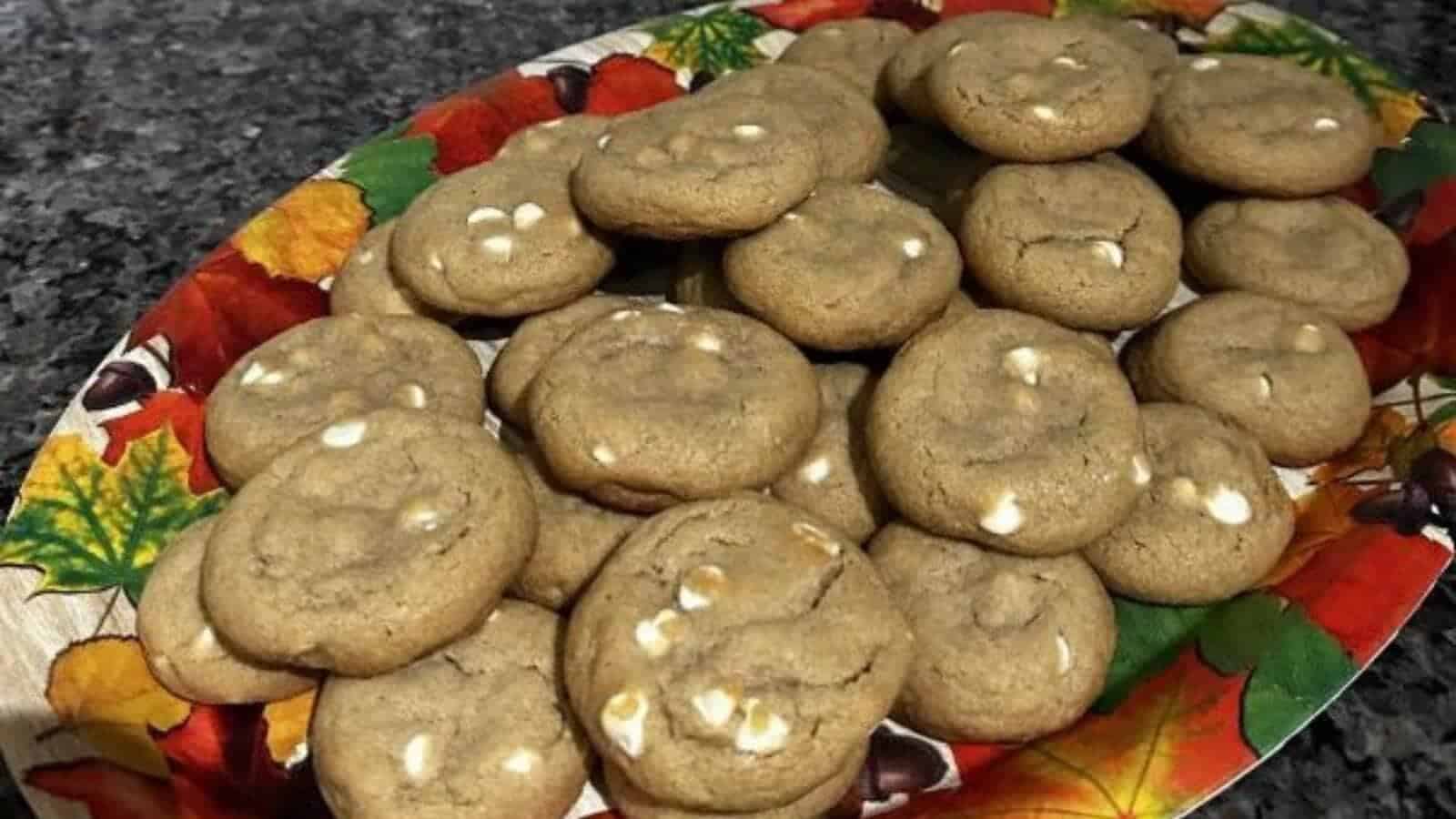 A plate of cookies with white chocolate chips, inspired by classic pumpkin recipes, is arranged on a decorative autumn-themed platter, set on a granite countertop.