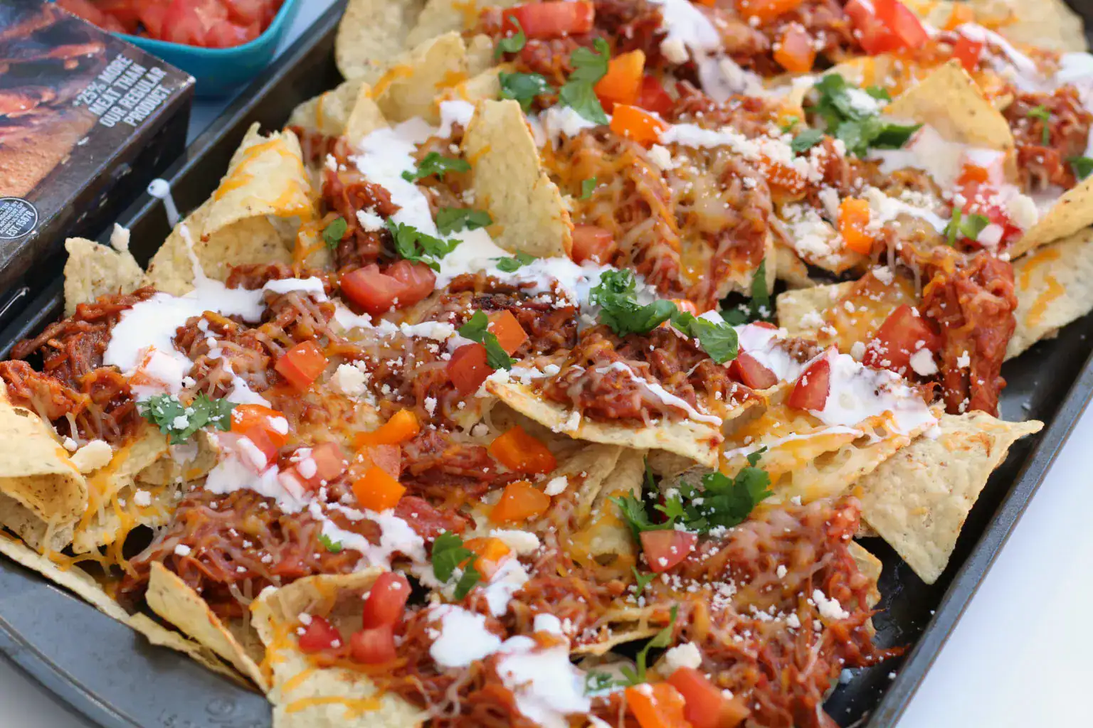 Tray of nachos topped with shredded meat, melted cheese, chopped tomatoes, diced bell peppers, cilantro, and a drizzle of white sauce.