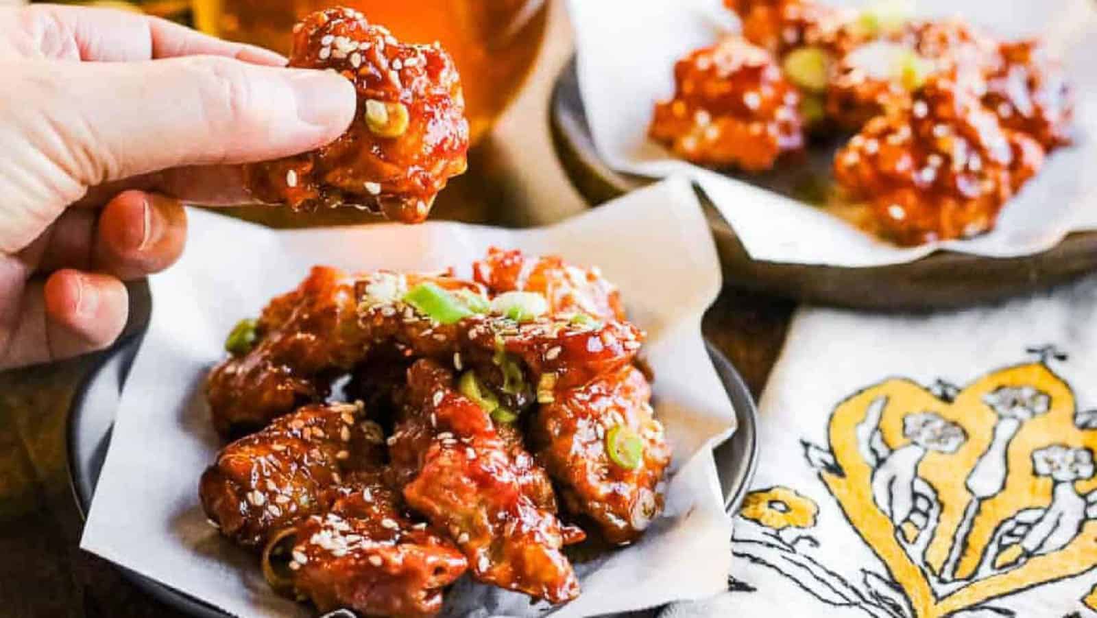 A hand holds a piece of saucy, sesame-topped fried chicken above a plate of similar pieces, garnished with green onions, next to a decorative napkin.