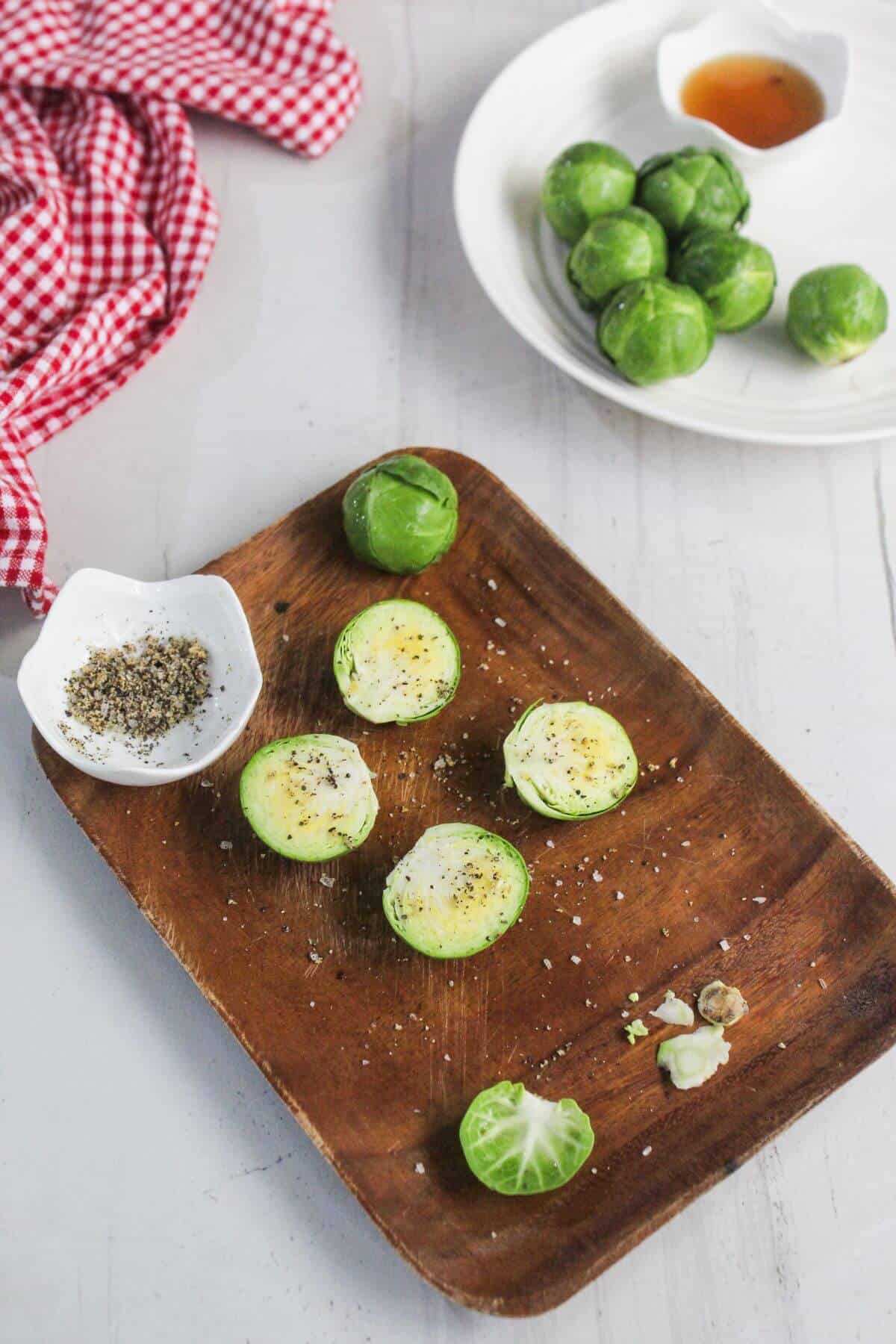Halved Brussels sprouts seasoned with salt and pepper on a wooden tray, whole Brussels sprouts and sauces in small bowls nearby on a white surface.