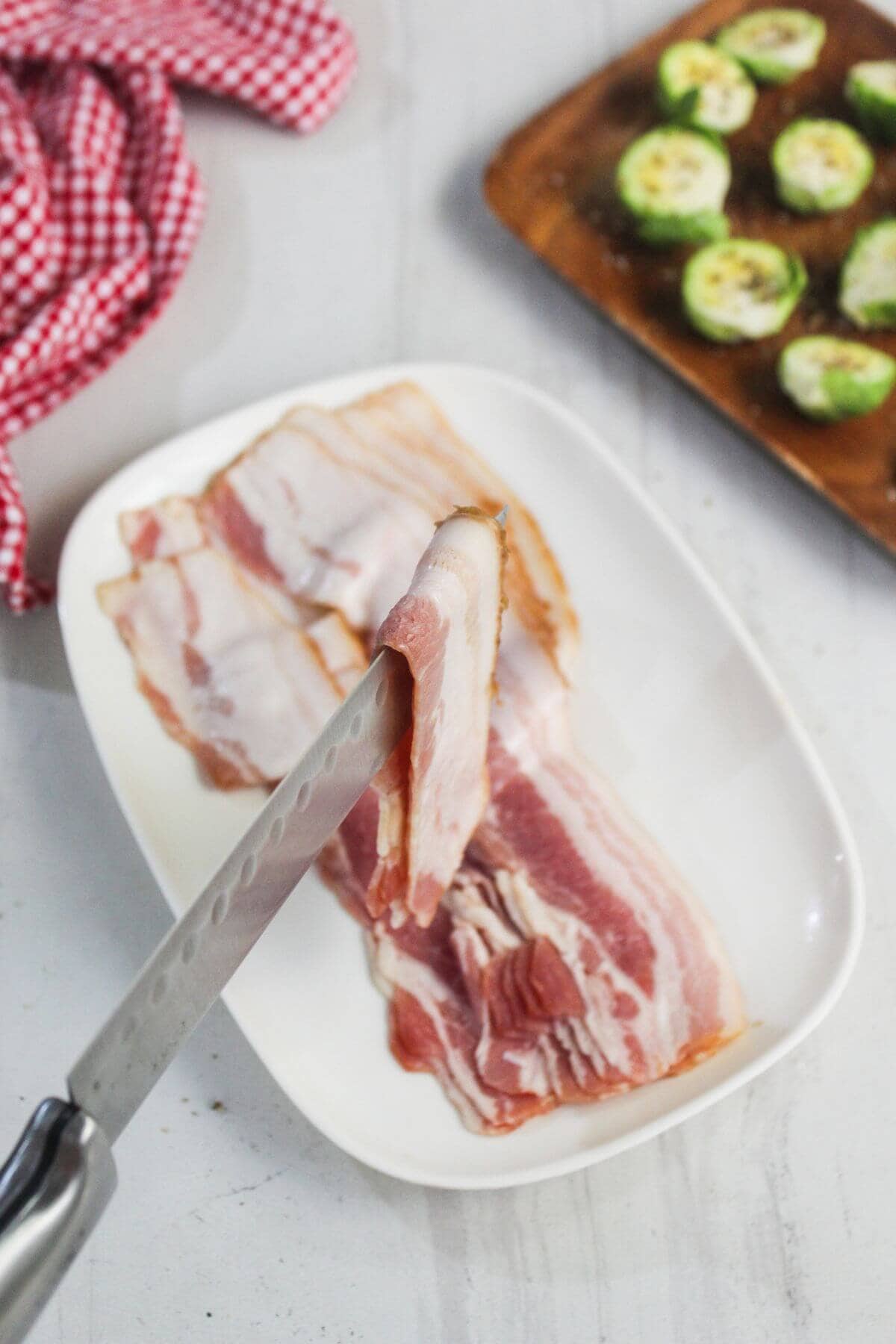 A knife slices raw bacon on a white plate, with a red checkered cloth and a tray of sliced Brussels sprouts in the background.
