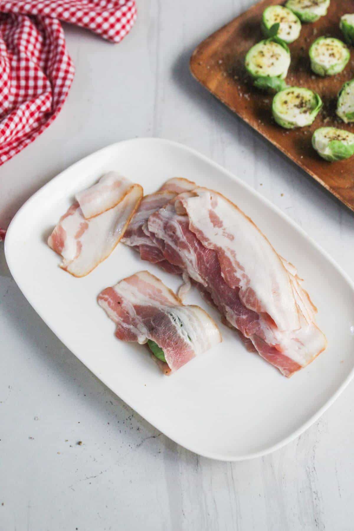 Slices of raw bacon on a white plate, with a red checkered cloth and a tray of seasoned Brussels sprouts in the background.