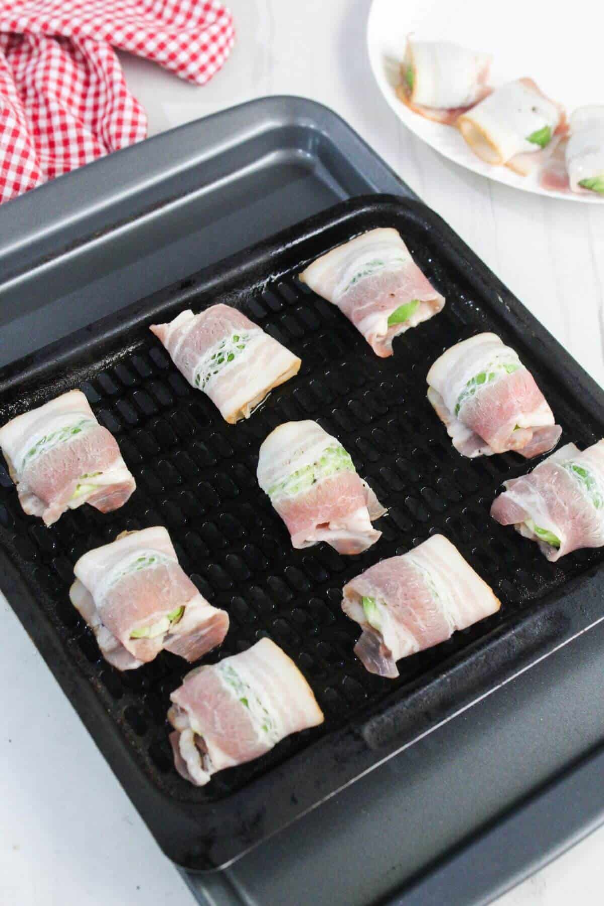 Bacon-wrapped cheese and jalapeño pieces arranged on a grill pan, ready to be cooked, with more on a plate in the background.