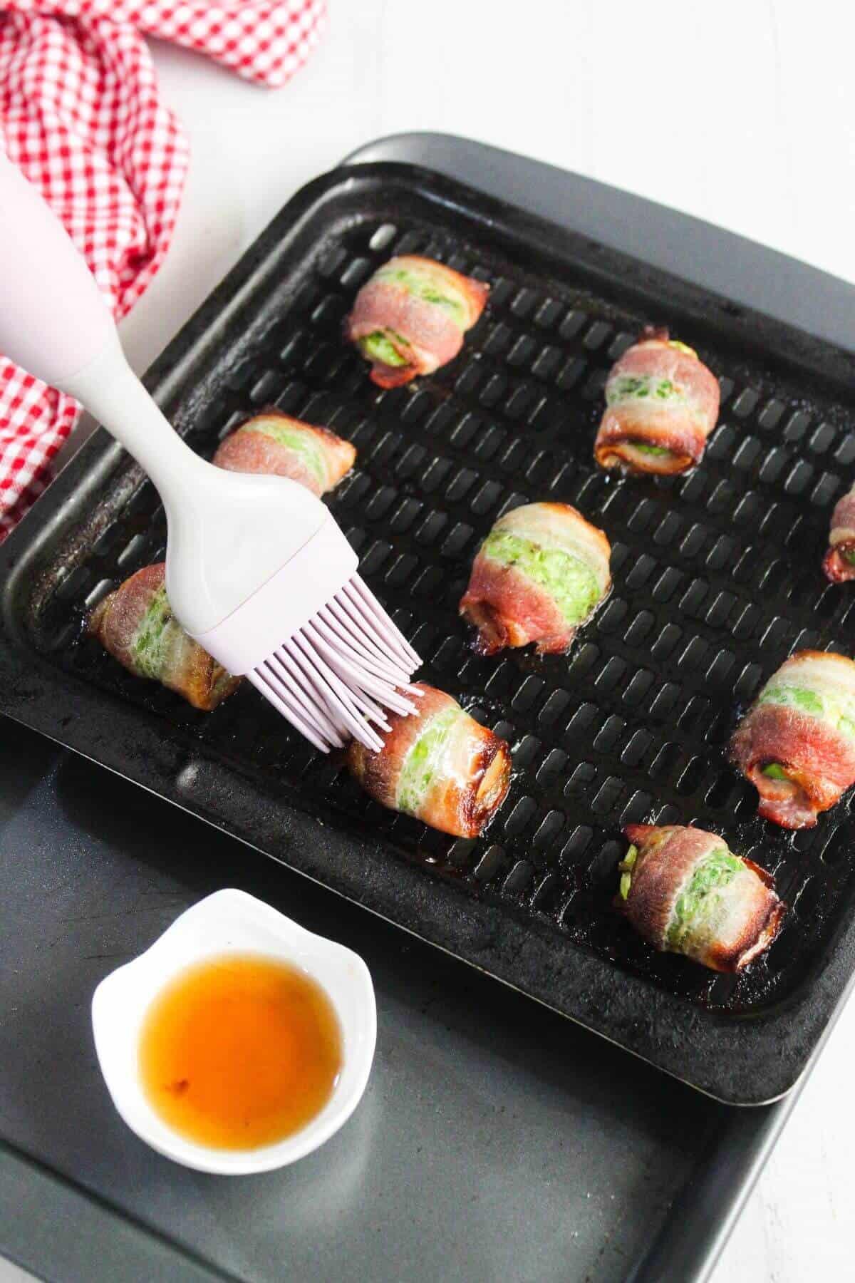 A silicone brush applies sauce to bacon-wrapped jalapeño poppers on a black grill pan, with a small bowl of sauce nearby.