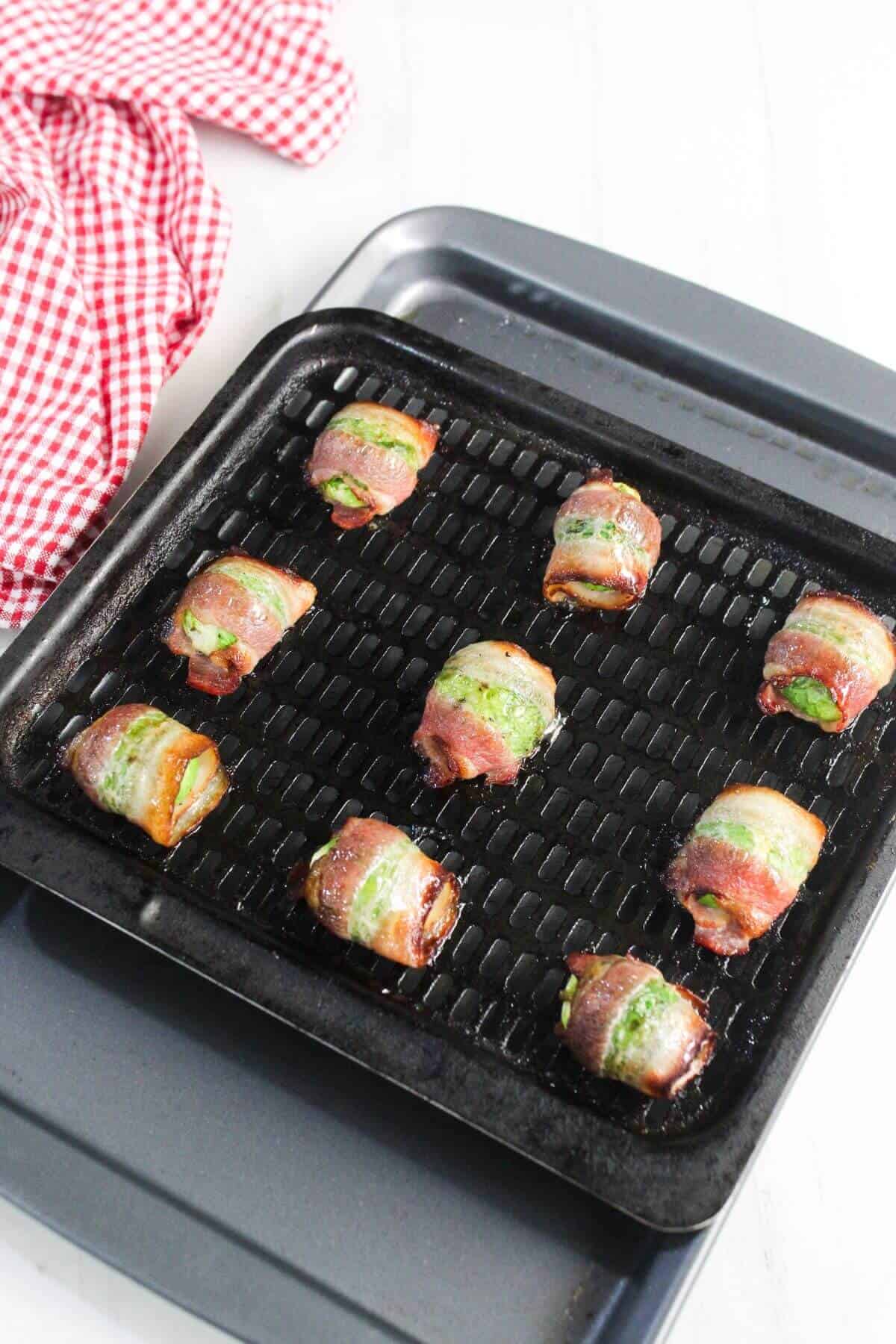Nine pieces of bacon-wrapped food are arranged on a black grill tray, with a red and white checkered cloth nearby on a white surface.