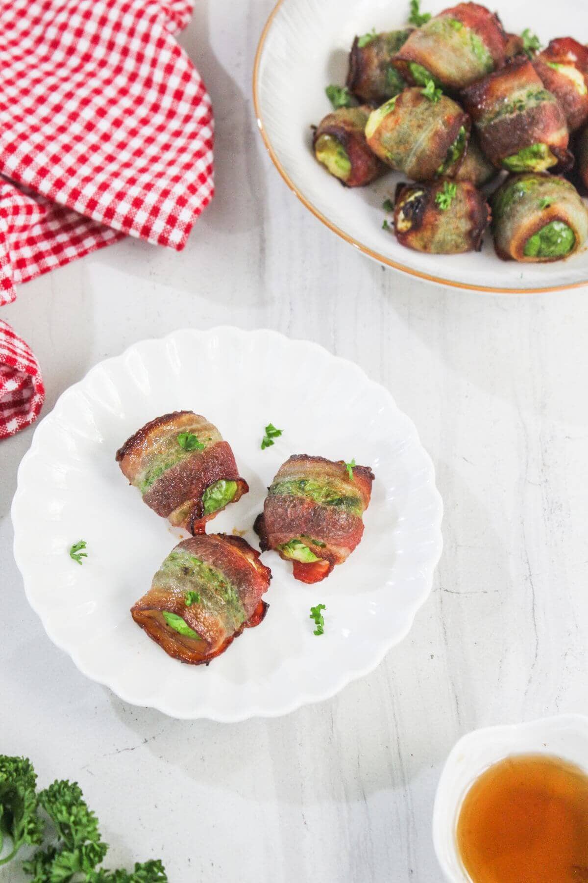 Three bacon-wrapped jalapeño poppers on a white plate, with more poppers in a bowl, a red checkered cloth, and a small bowl of sauce nearby.