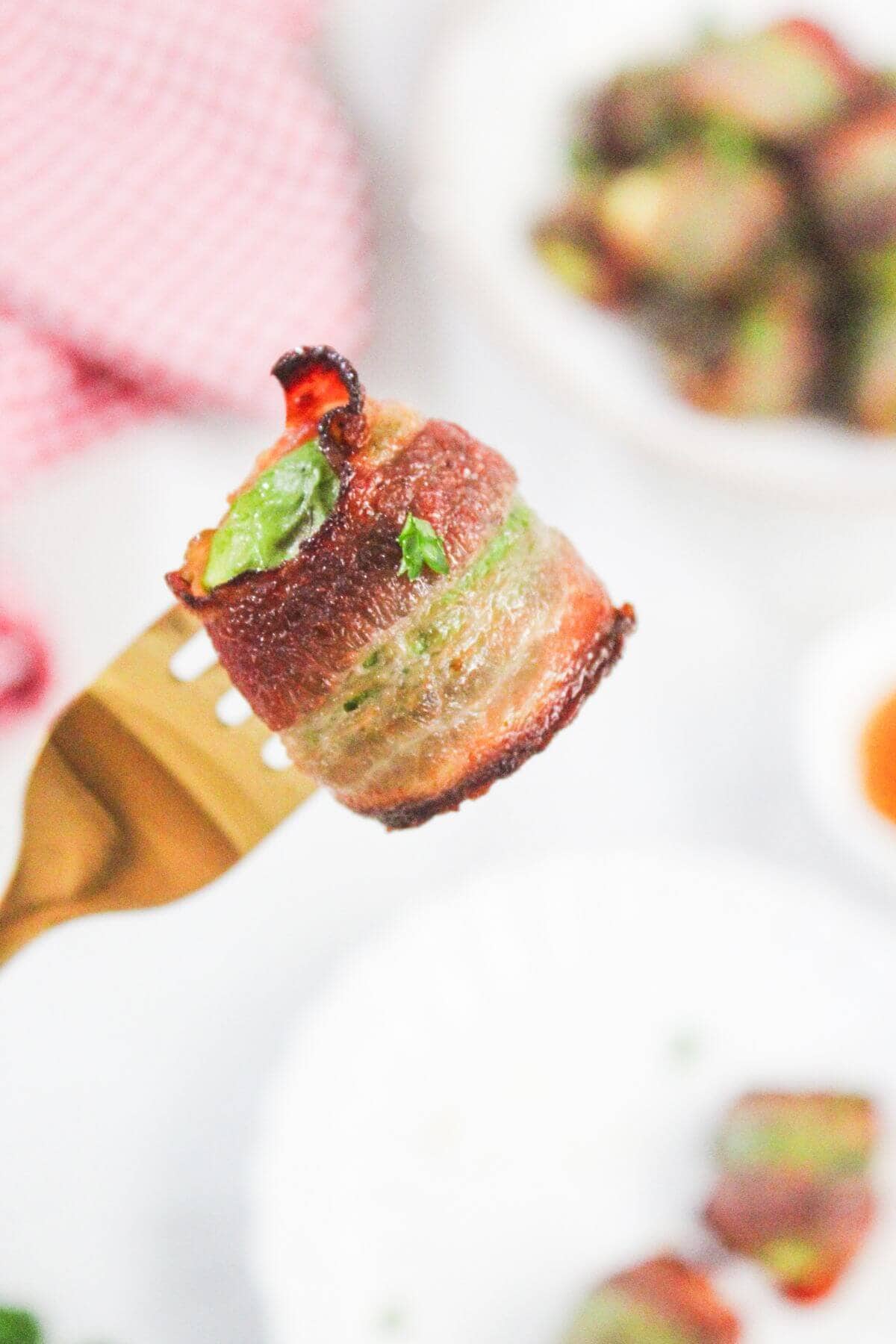 A close-up of a bacon-wrapped jalapeño bite held on a fork, with more bites blurred in the background.