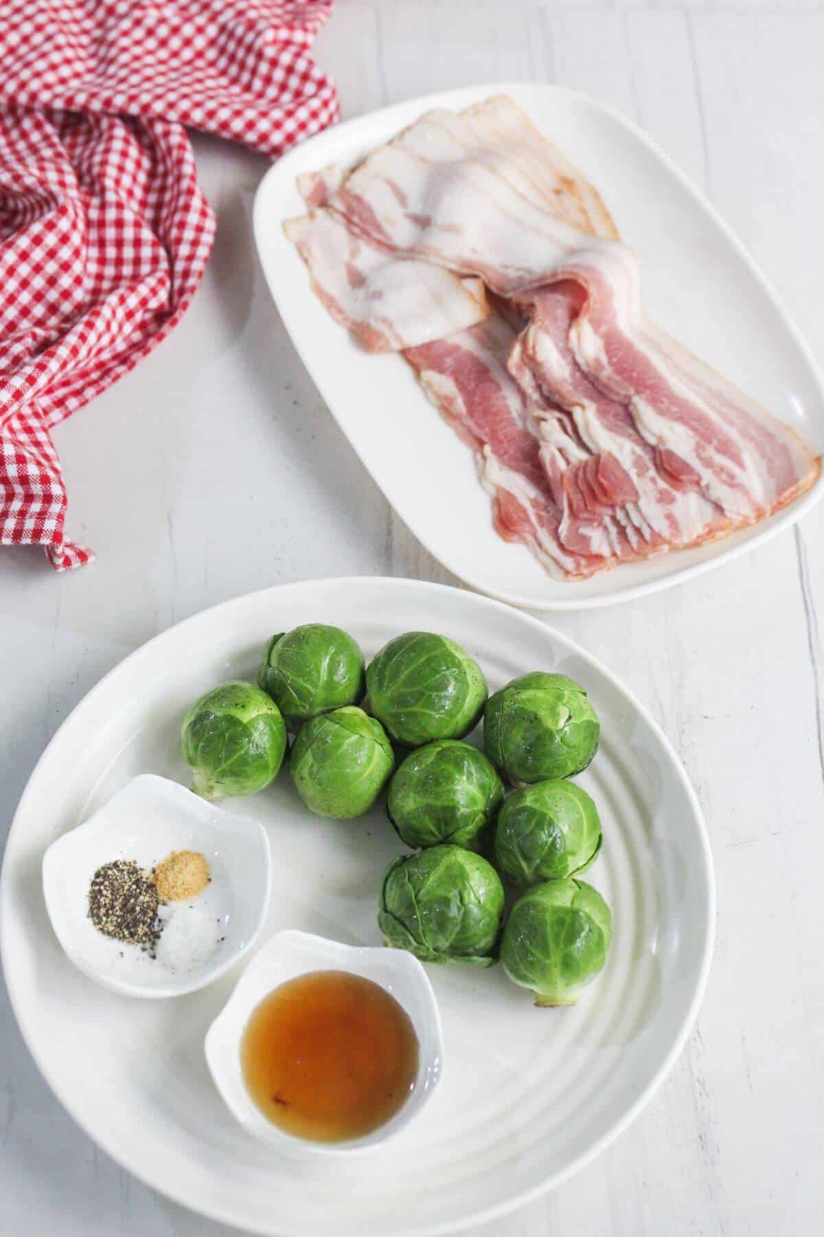 A plate with raw Brussels sprouts, seasonings, and a small bowl of liquid next to another plate with raw bacon slices on a white surface, with a red checkered cloth nearby.