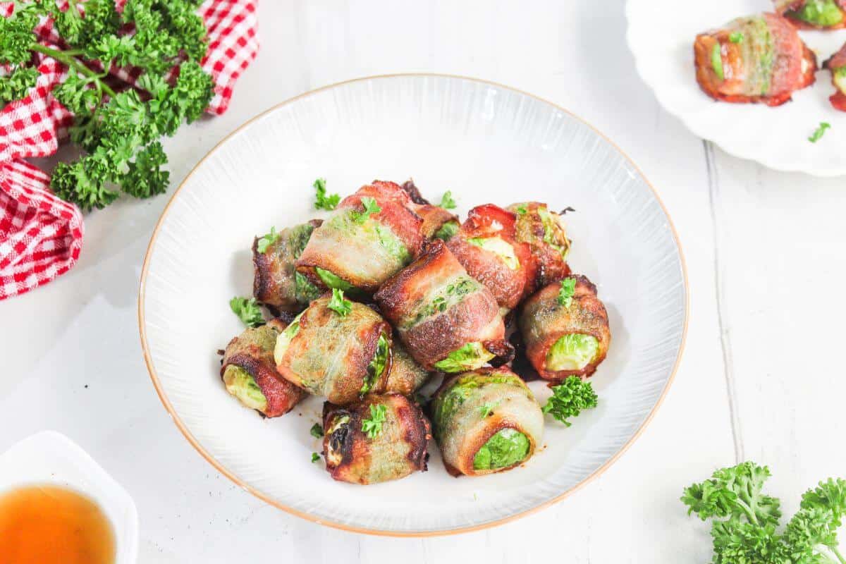 A plate of bacon-wrapped Brussels sprouts garnished with chopped parsley, with a side of dipping sauce and a red checked napkin nearby.