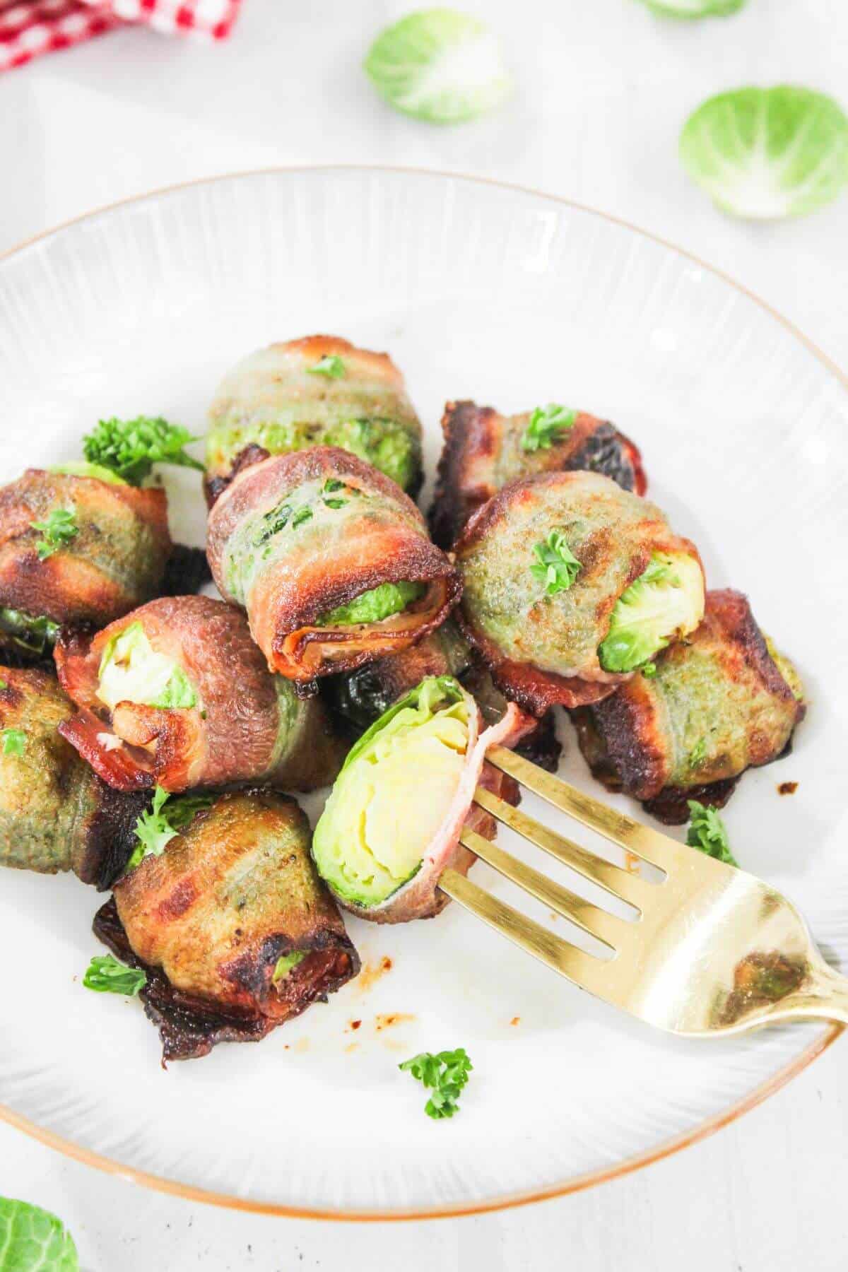 A plate of bacon-wrapped Brussels sprouts garnished with chopped parsley, with a fork holding one piece on a white plate.
