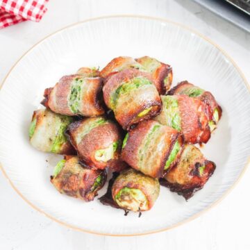 A plate of bacon-wrapped jalapeño poppers filled with cream cheese, arranged in a white dish on a light surface.