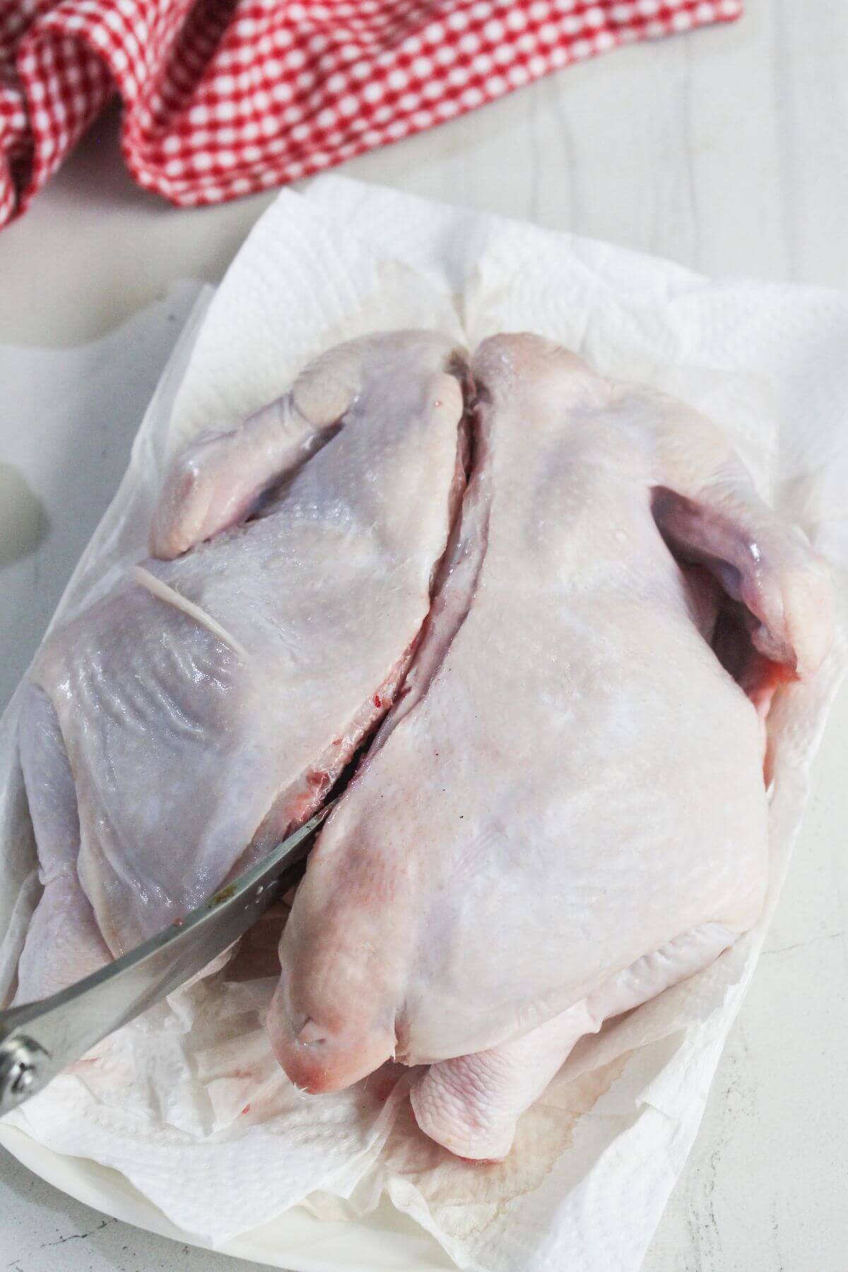 A raw whole chicken is being butterflied with a knife on a paper towel-lined surface, next to a red and white checkered cloth.