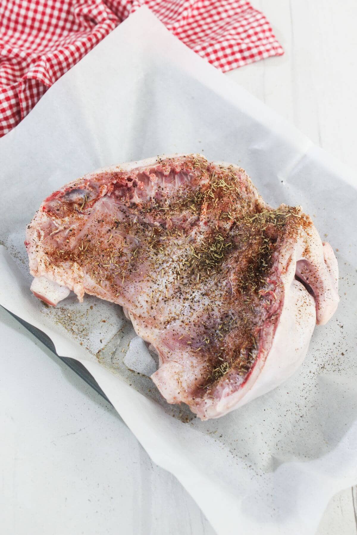 Raw turkey breast with skin, sprinkled with herbs and spices, placed on parchment paper in a baking dish; red checked cloth in background.