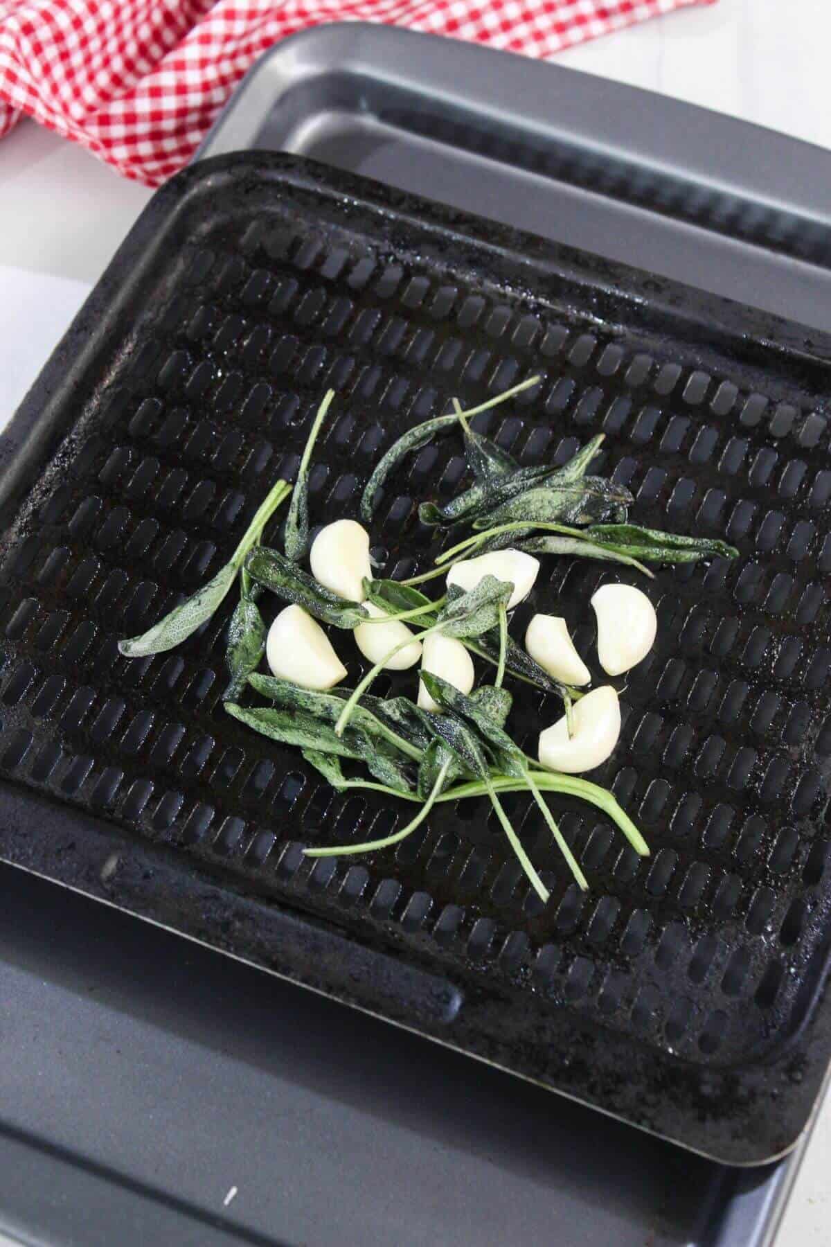 Whole garlic cloves and fresh sage leaves arranged on a black grill pan, ready to be roasted; a red and white checkered cloth is in the background.
