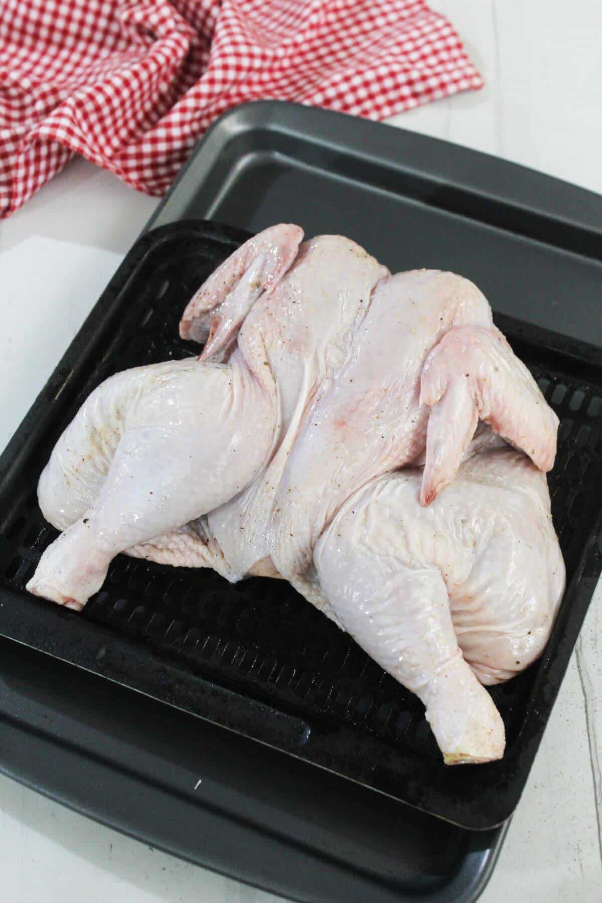 Raw spatchcocked chicken placed on a black roasting tray, with a red and white checkered cloth in the background.