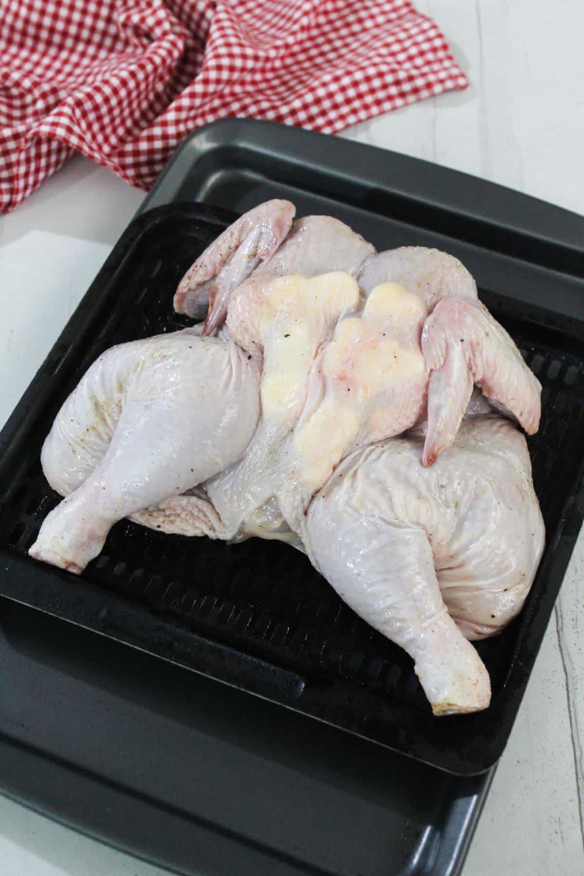 A raw spatchcocked chicken is placed on a black roasting tray, with a red and white checkered cloth in the background.