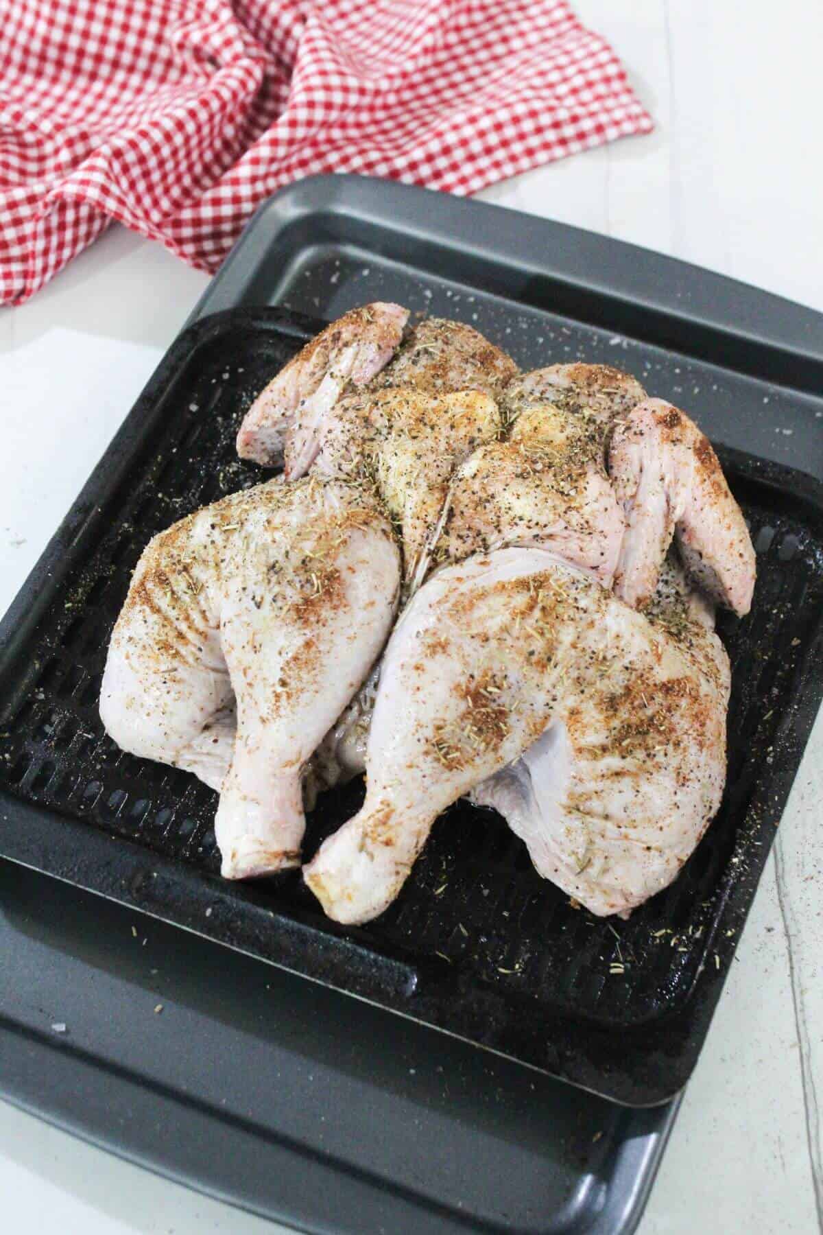 A raw, spatchcocked chicken seasoned with spices sits on a black roasting pan, with a red and white checkered cloth in the background.