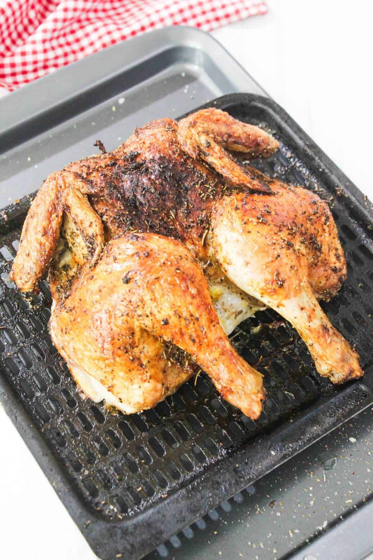 A cooked, seasoned spatchcock chicken rests on a black roasting rack, with a red and white checkered cloth in the background.