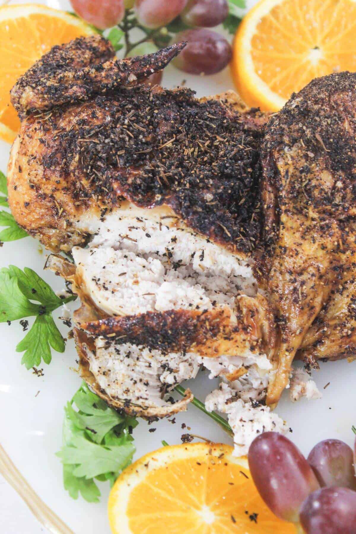 Roasted chicken with herb seasoning is sliced and served on a platter, garnished with orange slices, parsley, and red grapes.