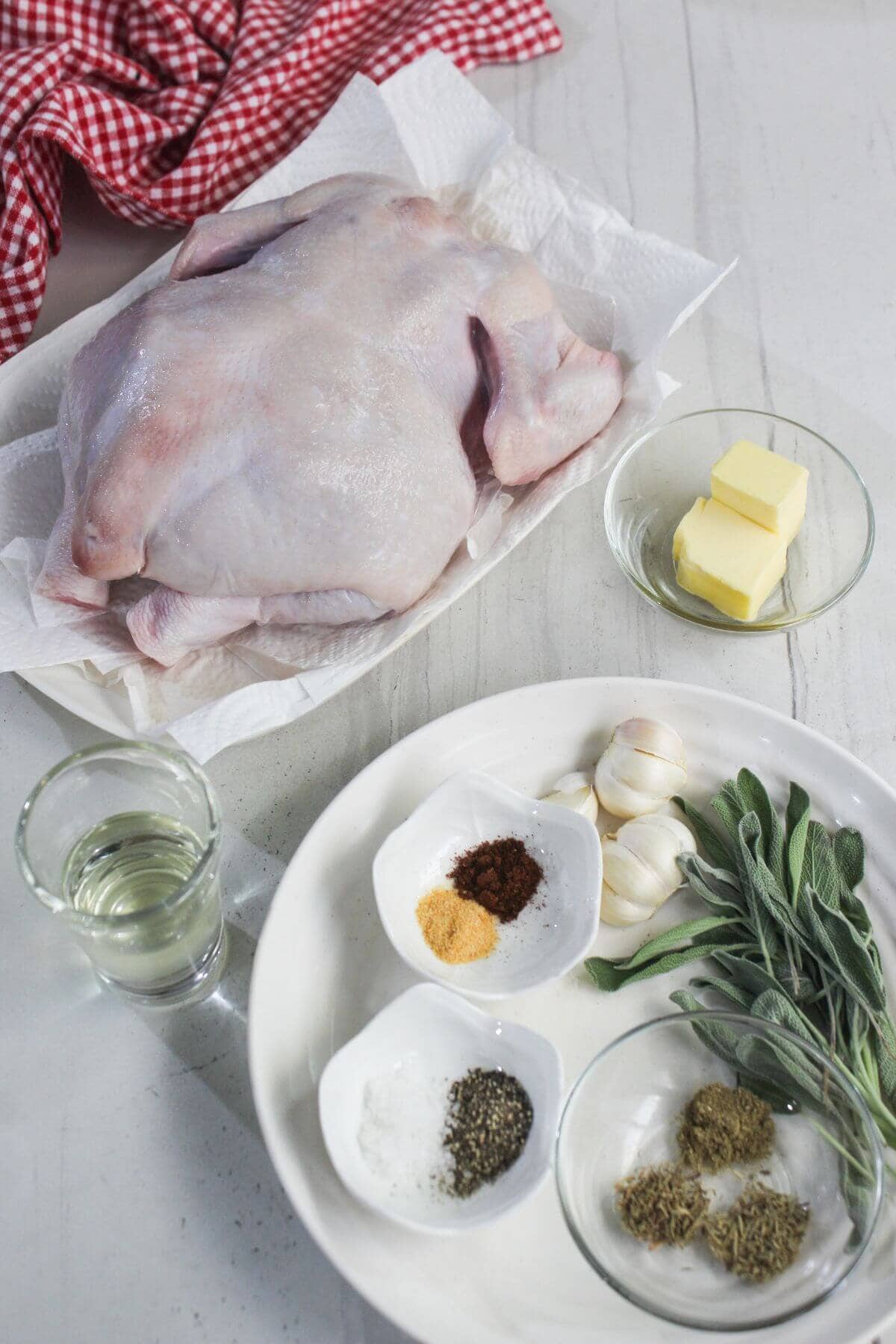 Uncooked whole chicken on a plate with butter, garlic, fresh sage, spices, herbs, and a glass of liquid on a white table.