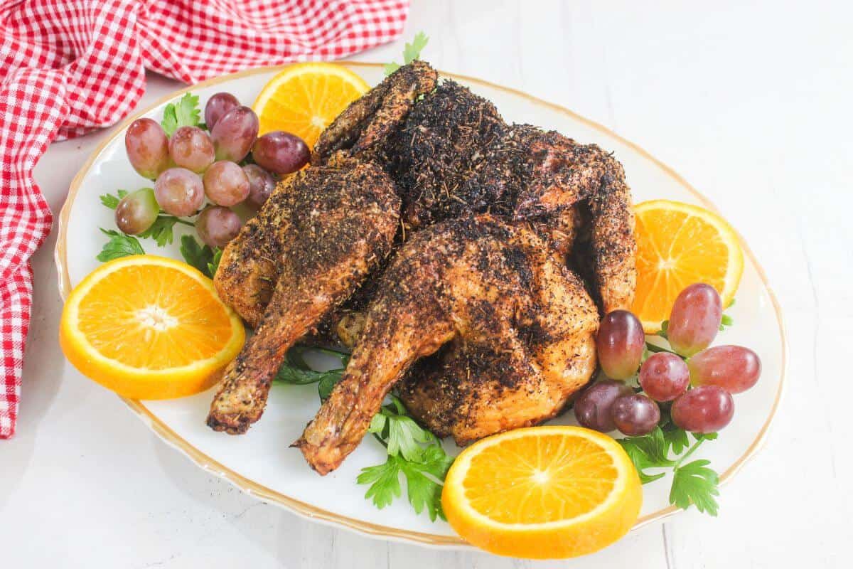 A roasted, seasoned chicken is served on a white platter with orange slices, red grapes, fresh parsley, and a red gingham cloth in the background.