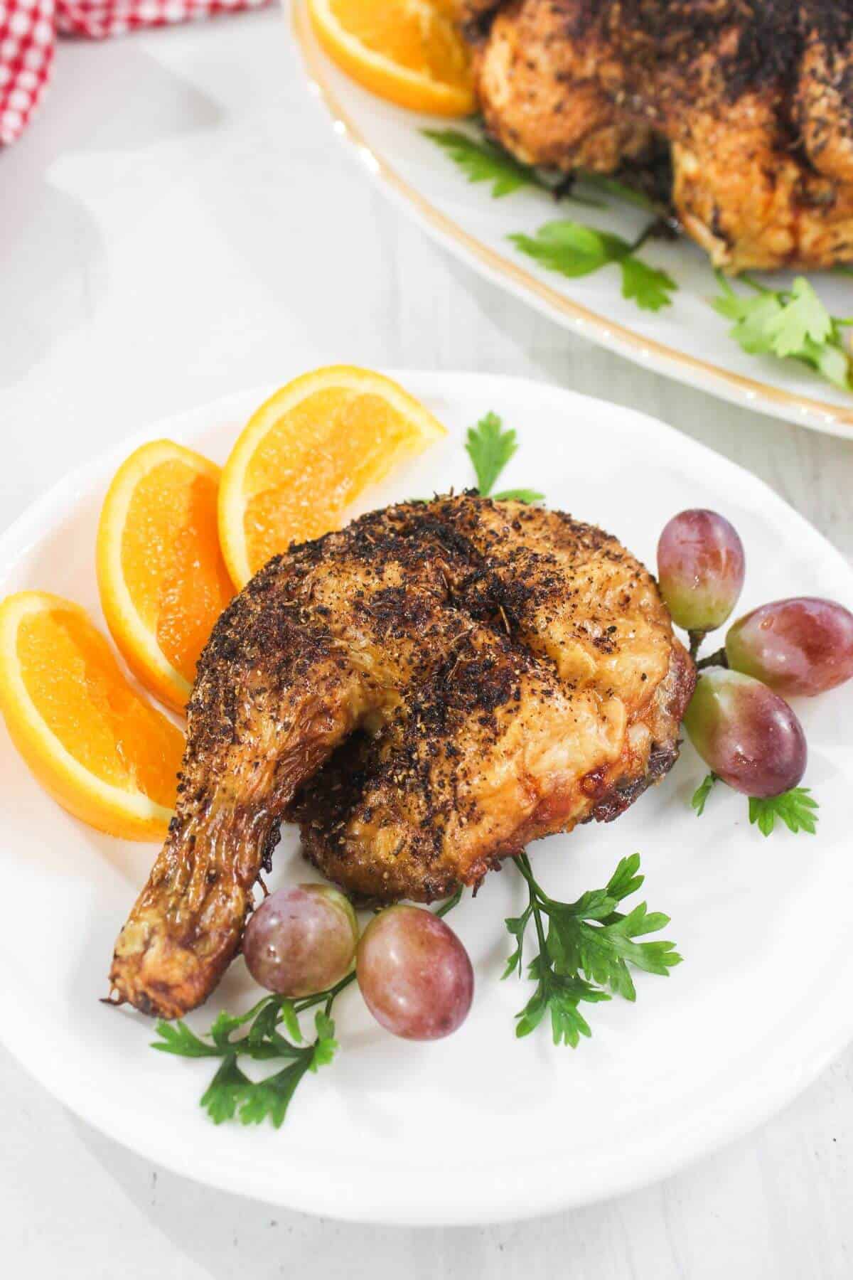 A cooked chicken leg quarter is served on a white plate with orange slices, red grapes, and fresh parsley as garnish.