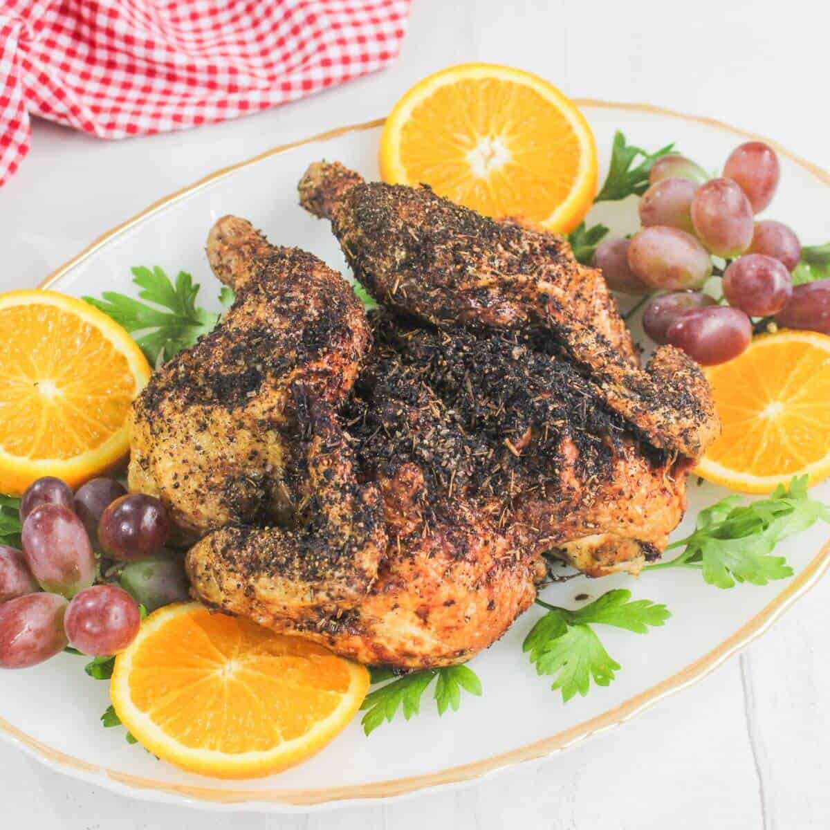 Roast chicken seasoned with herbs, garnished with orange slices, red grapes, and parsley on a white platter.