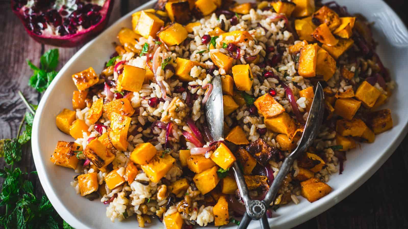 A white platter of rice salad topped with roasted butternut squash cubes, red onions, herbs, and walnuts, with serving utensils placed on the dish.