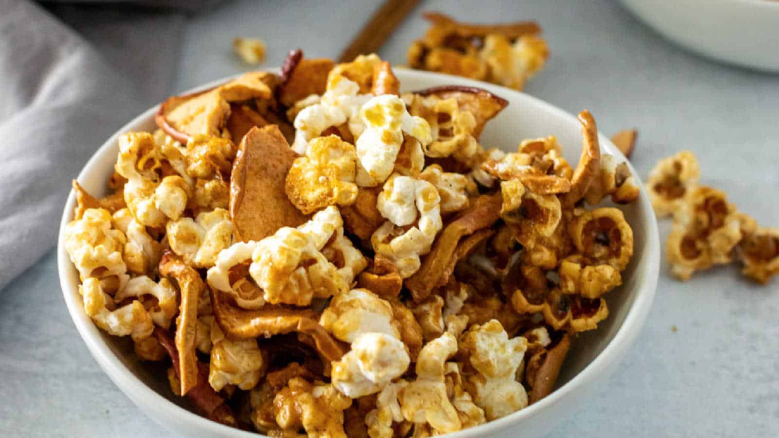 A white bowl filled with caramel popcorn and pieces of dried apple, with some pieces scattered on the surface nearby.