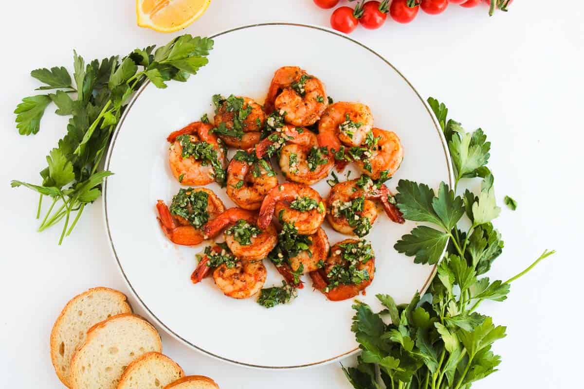 A plate of cooked shrimp garnished with chopped herbs, surrounded by fresh parsley, tomato vine, lemon wedge, and sliced baguette on a white background.