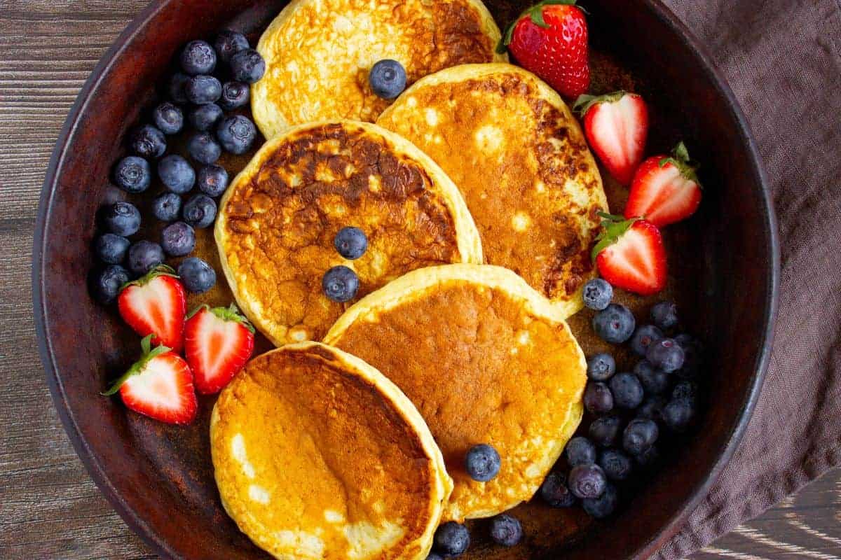 A plate with five pancakes, fresh blueberries, and halved strawberries arranged around the edge.