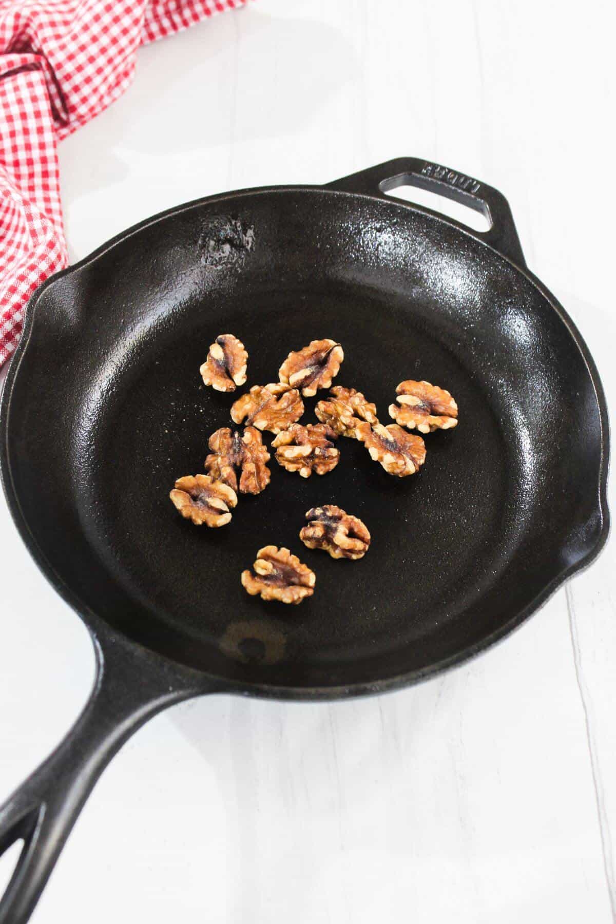 A few walnut halves in a black cast iron skillet on a white surface, with a red and white checkered cloth nearby.