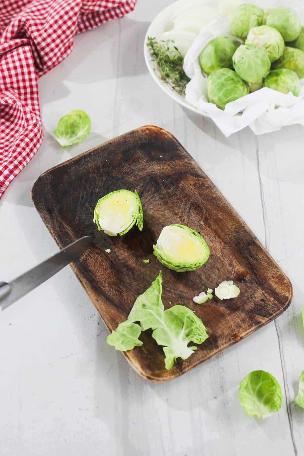 A halved Brussels sprout on a wooden cutting board with a knife, loose leaves, and a bowl of whole Brussels sprouts nearby.