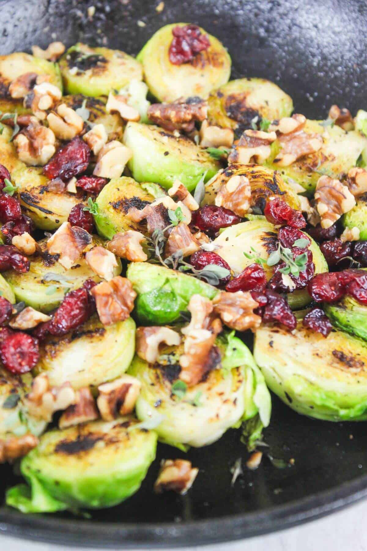 Sautéed Brussels sprouts in a pan, topped with chopped walnuts, dried cranberries, and fresh herbs.