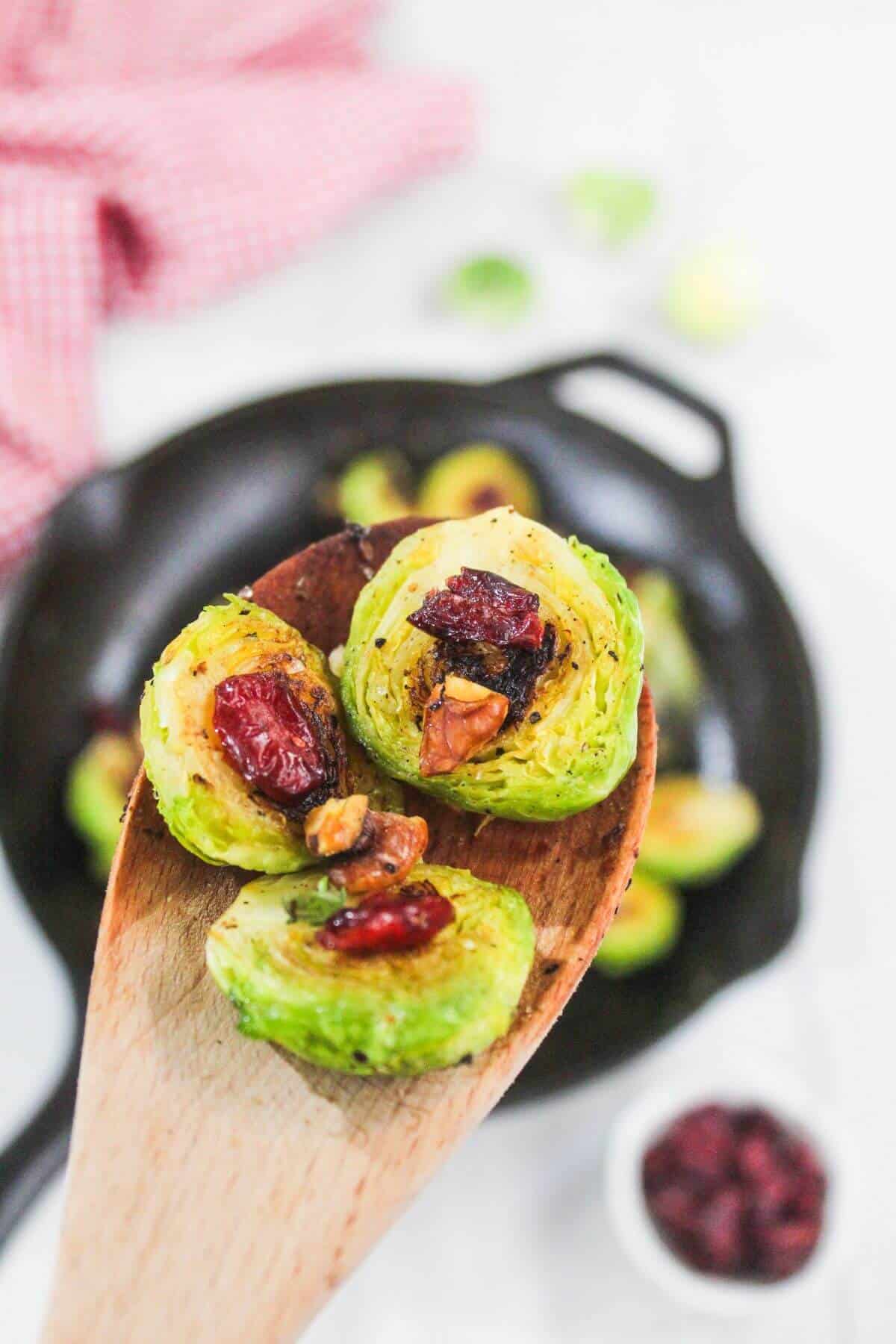 A wooden spoon holds roasted Brussels sprouts topped with dried cranberries and pecans, with a skillet and red cloth in the background.