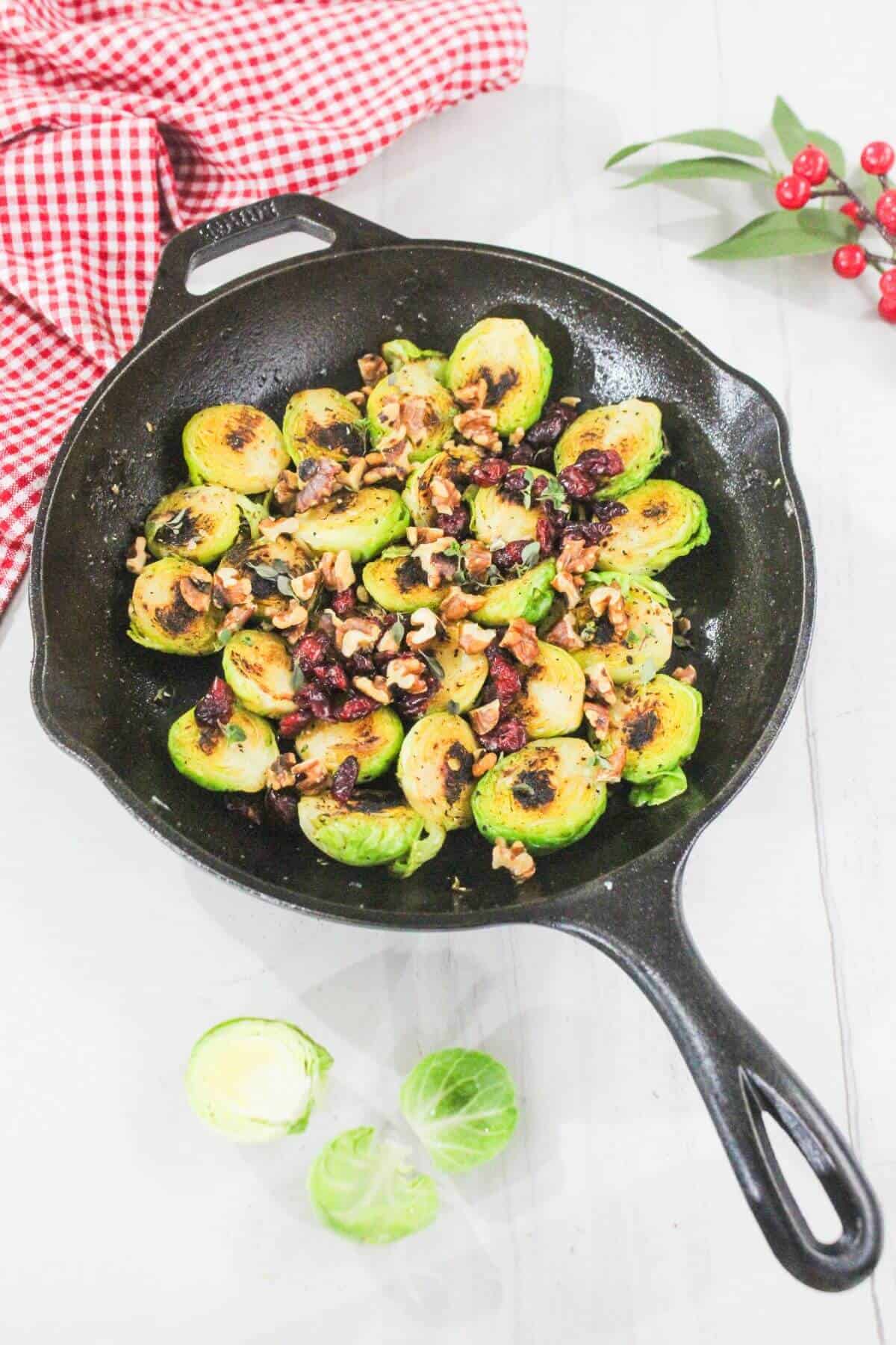A cast iron skillet with roasted Brussels sprouts, walnuts, and dried cranberries on a light surface, with a red checkered cloth and loose Brussels sprouts nearby.